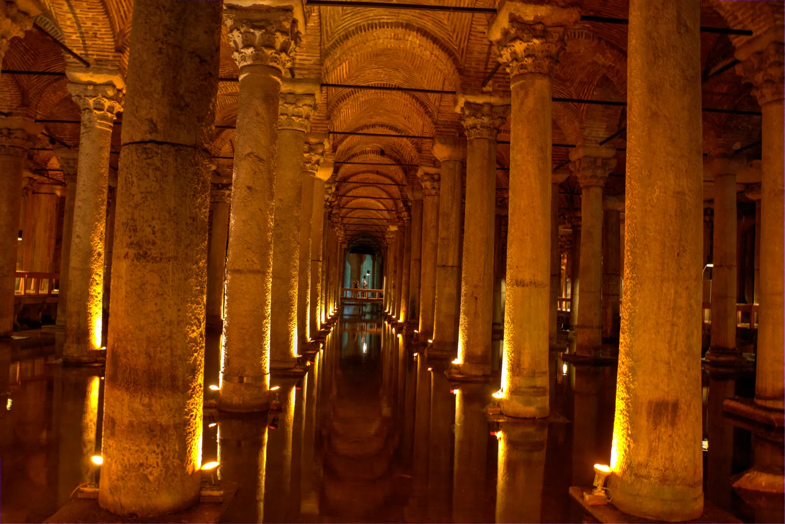 The interior of the Basilica Cistern, rows of tall marble columns lit from their bases in amber light, receding into the dark under brick vaults, with shallow water reflecting them on the floor