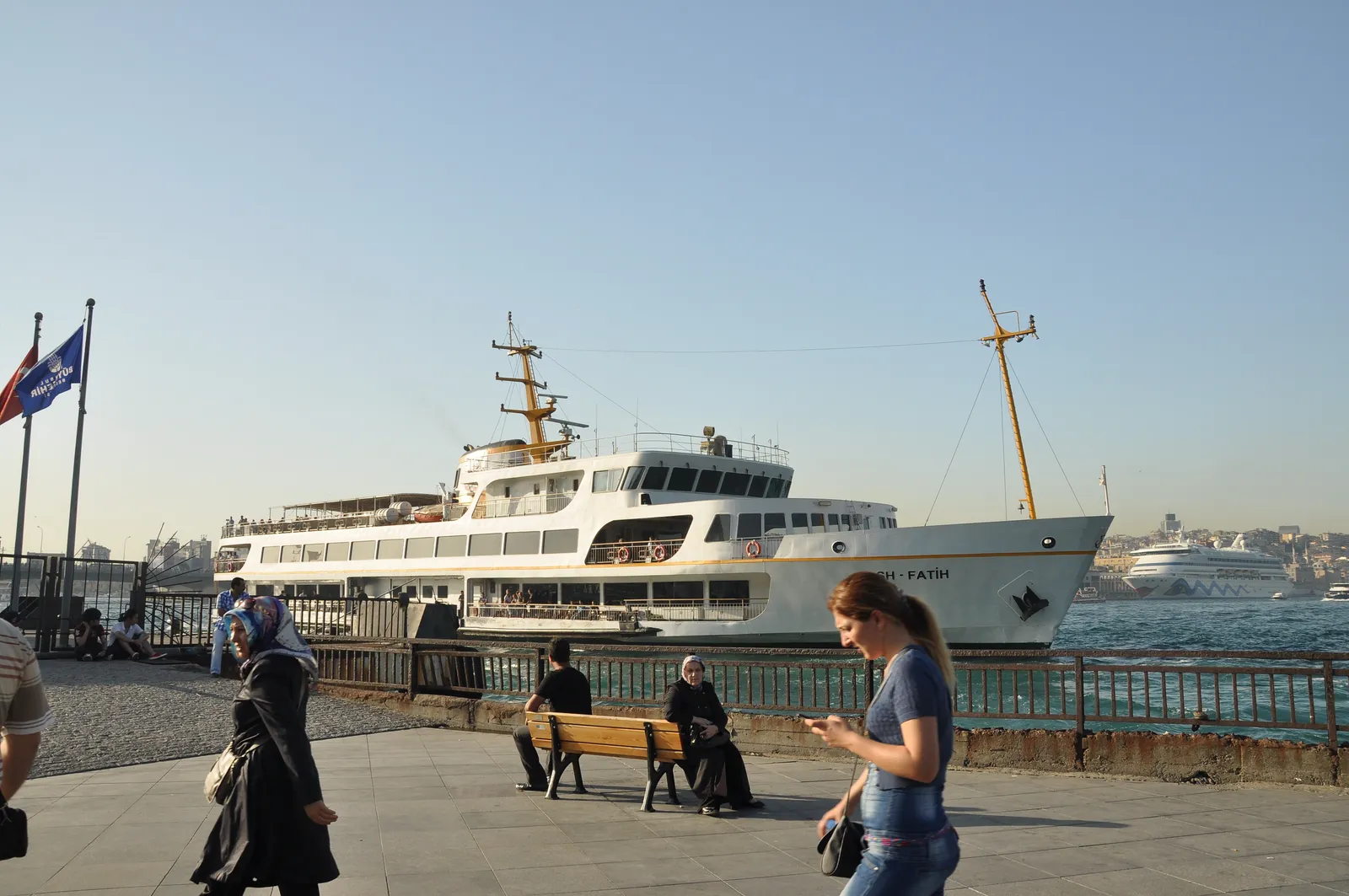 A white double-decker commuter ferry moored at the Eminönü pier in Istanbul, with people sitting and walking along the paved waterfront in front of it