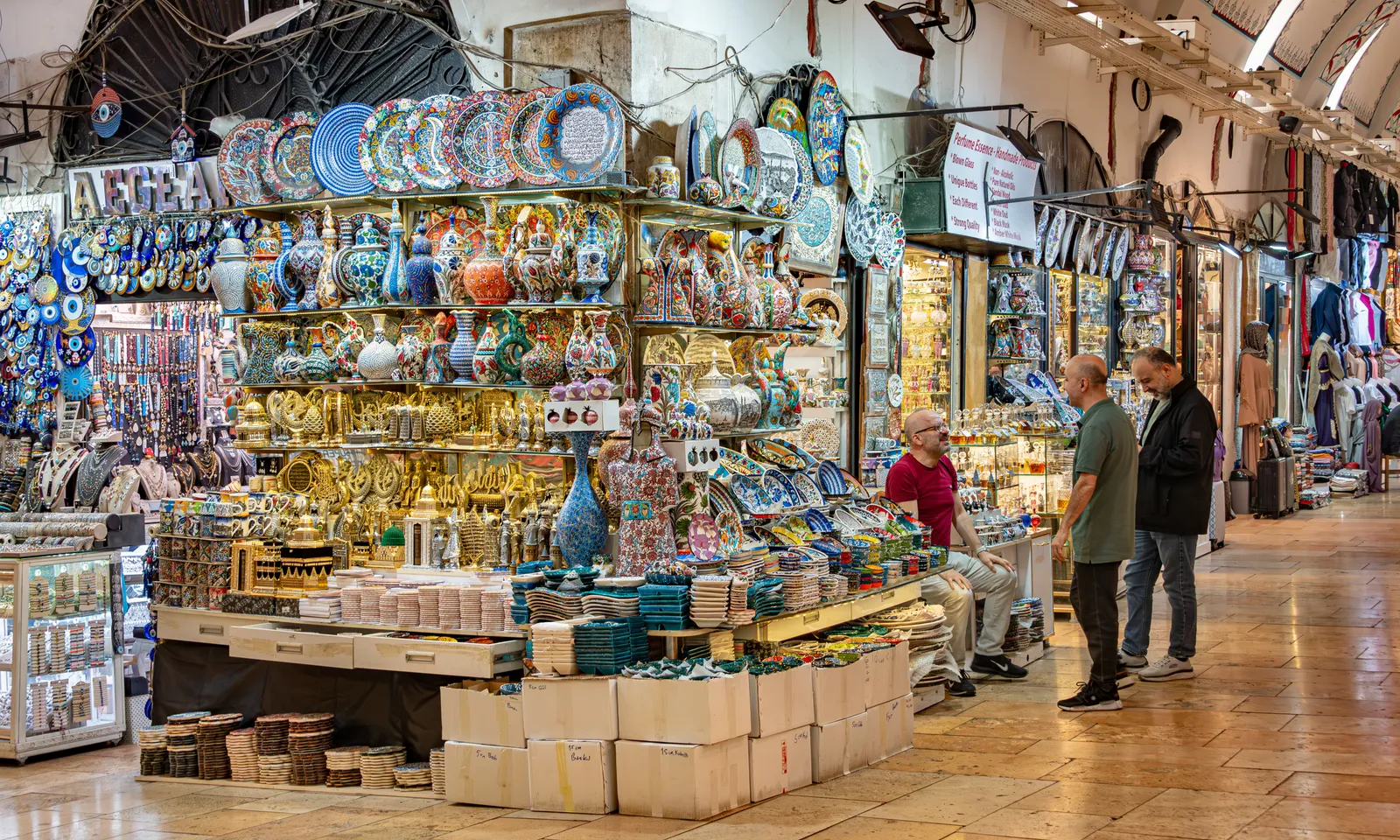 A covered street inside Istanbul's Grand Bazaar, stalls stacked to the vaulted ceiling with painted ceramic plates, bowls and brass lamps, and a few shoppers standing in the aisle