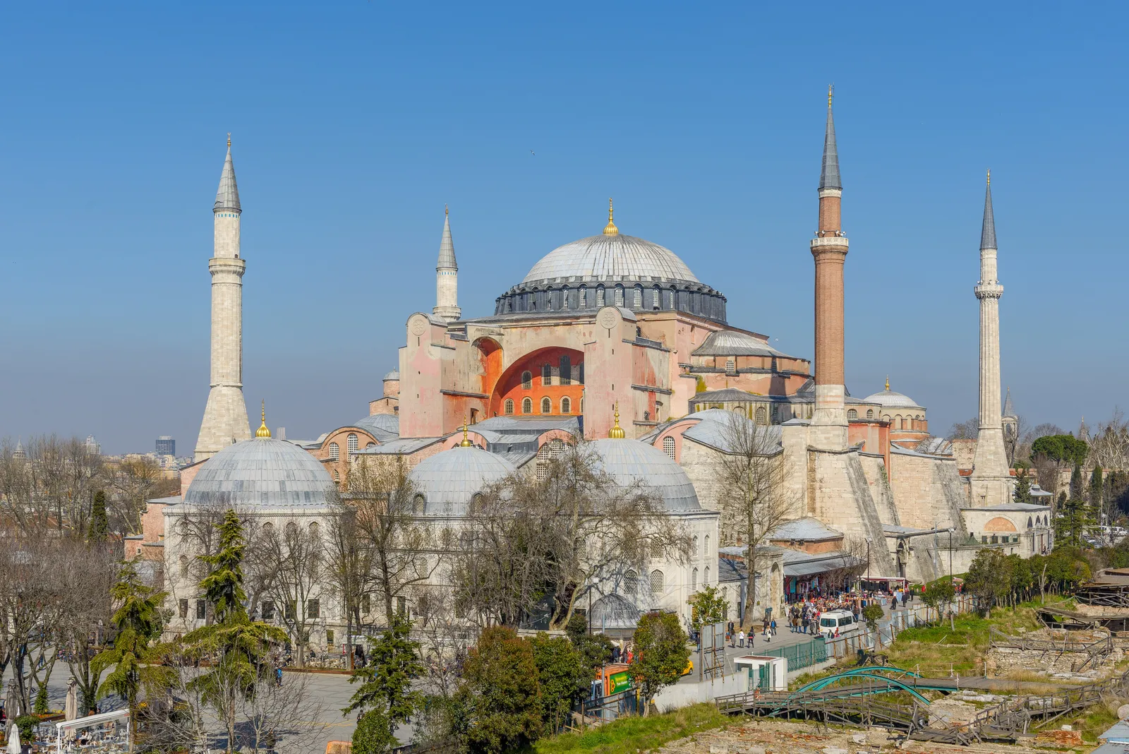 Hagia Sophia in daylight, its grey central dome ringed by half-domes above pale stone walls with red-brown buttresses, and four slender minarets rising around it against a clear blue sky
