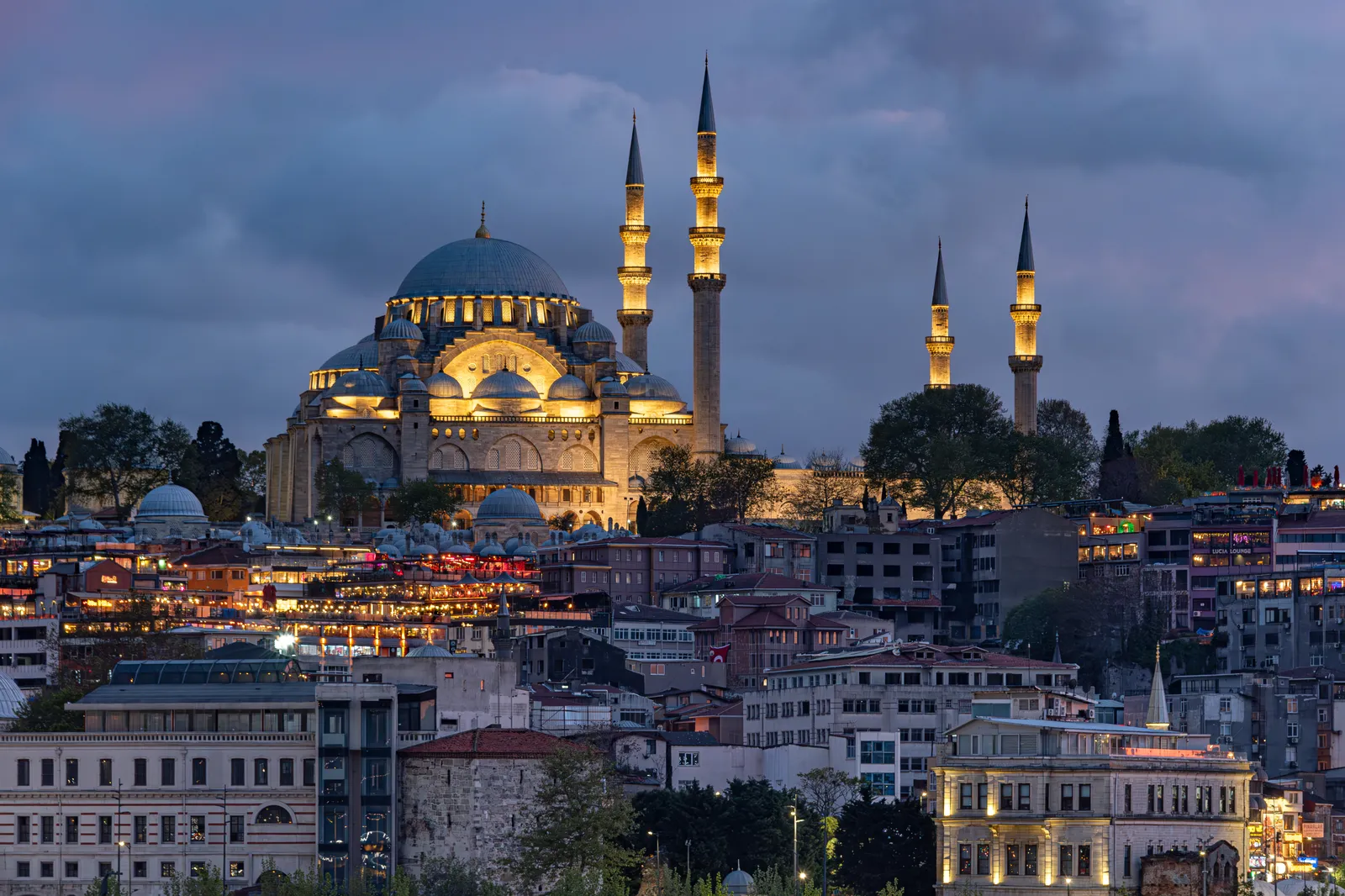 Süleymaniye Mosque lit at dusk on the ridge above the Golden Horn, its dome and four minarets picked out in gold light above the rooftops and lit windows of the streets below it