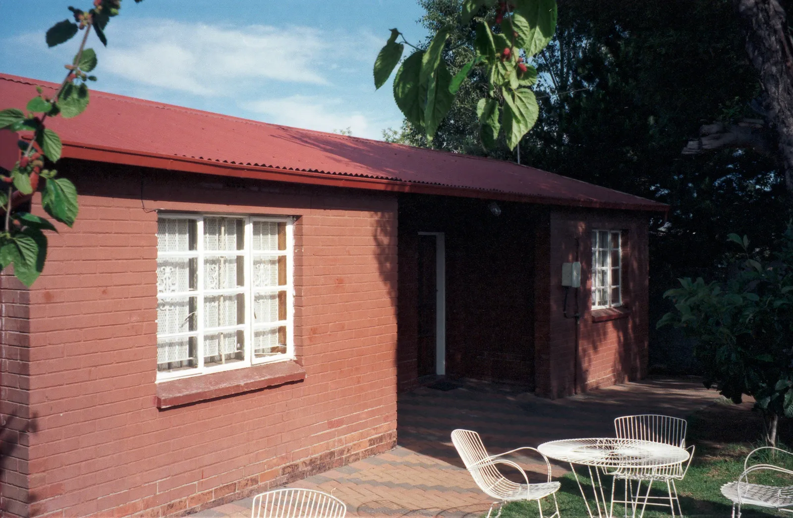 Nelson Mandela's former house on Vilakazi Street in Soweto, a small single-storey red brick building with a red corrugated iron roof, white window frames and a paved yard beside it