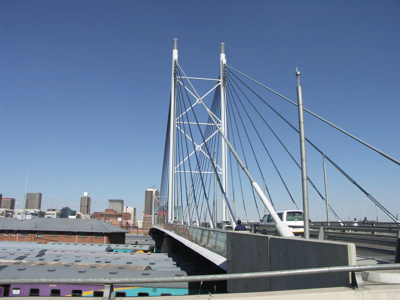 Nelson Mandela Bridge in Johannesburg seen from its deck, two white pylons carrying cables above the roadway, with the railway sheds and carriages of Park Station below and the city centre beyond
