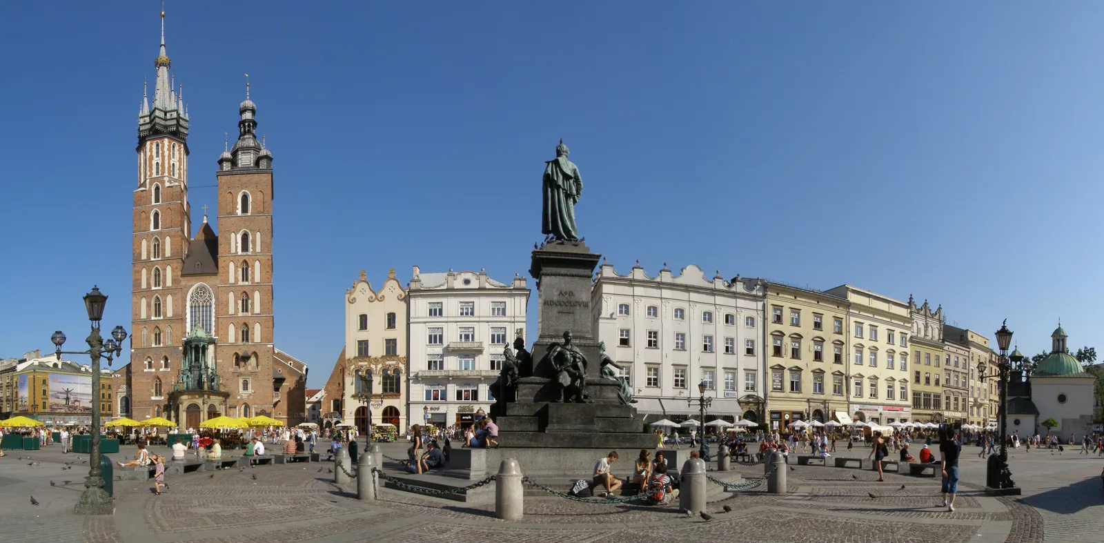 Kraków's Main Market Square on a clear day, the two unequal towers of St Mary's Basilica on the left, the Adam Mickiewicz monument in the middle and a row of pastel townhouses along the far side