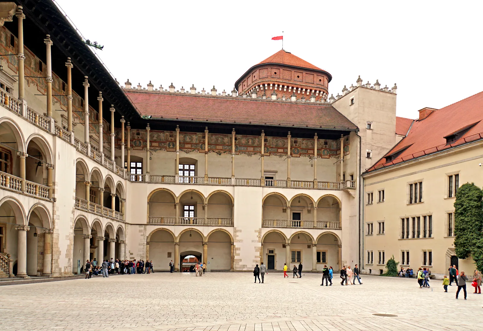 The arcaded courtyard inside Wawel Royal Castle, three tiers of Renaissance arches on slender columns running around a paved yard, with a red-roofed tower rising behind the far wing