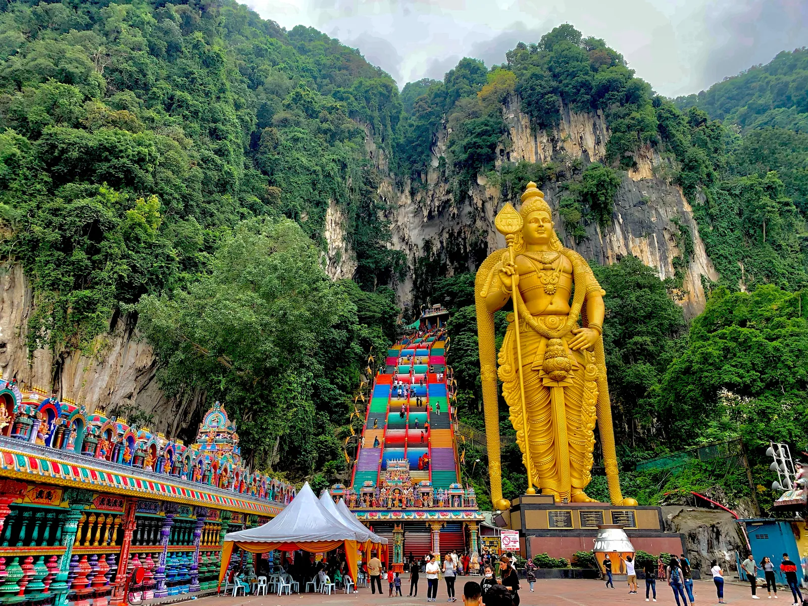 The rainbow-painted staircase at Batu Caves climbing the limestone cliff towards the cave mouth, with the gilded statue of Murugan standing beside it and forest on the rock face above