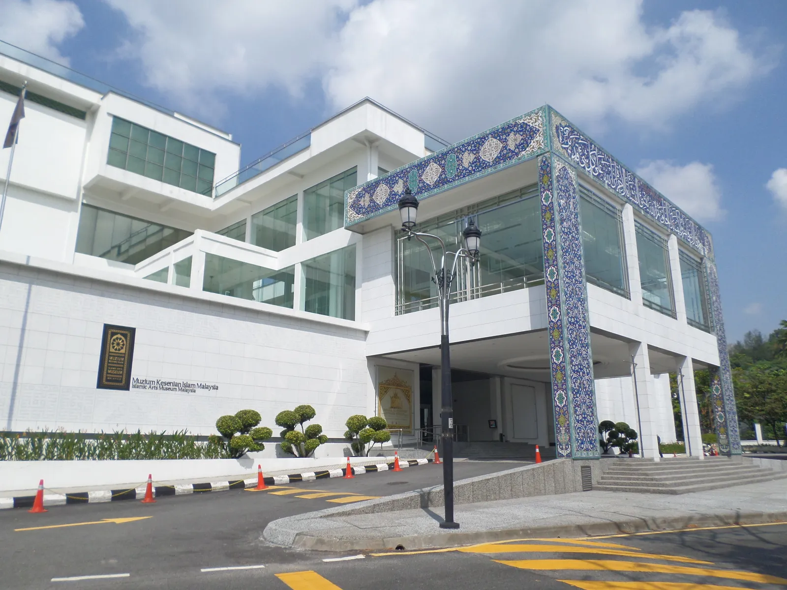 The white entrance block of the Islamic Arts Museum Malaysia in Kuala Lumpur, its porch faced in blue and white Iranian tilework, with clipped topiary along the forecourt