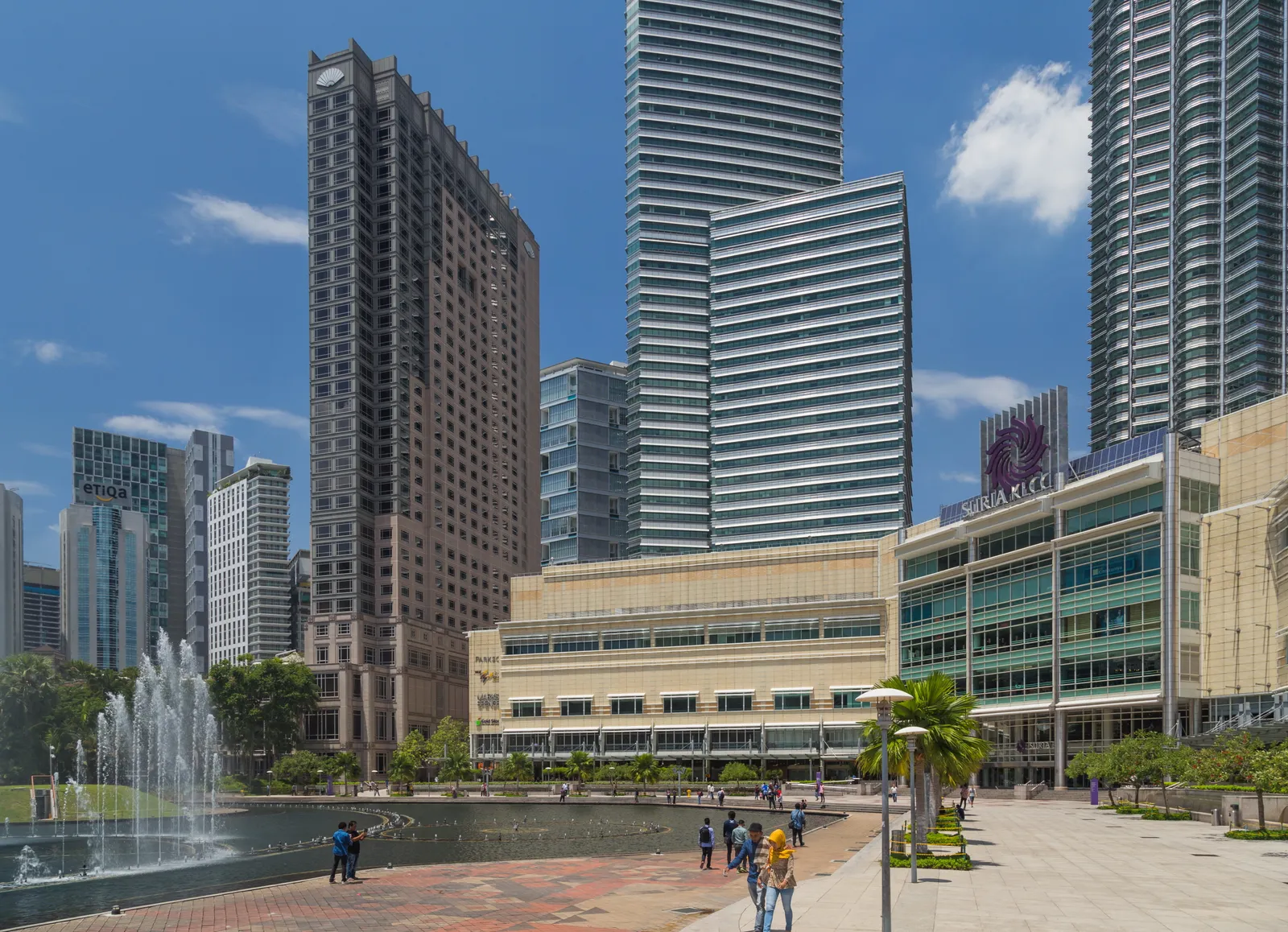 The esplanade at KLCC Park in Kuala Lumpur, water jets playing across the lake in the foreground and people crossing the paved terrace in front of the Suria KLCC shopping centre and surrounding office towers