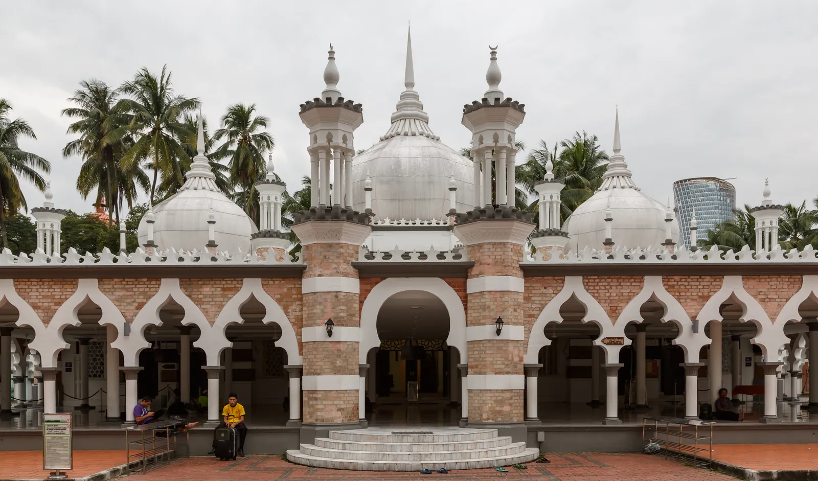 The front of Jamek Mosque in Kuala Lumpur, white onion domes and slender minarets above an arcade of pink and white banded brick arches, with palms behind the roofline