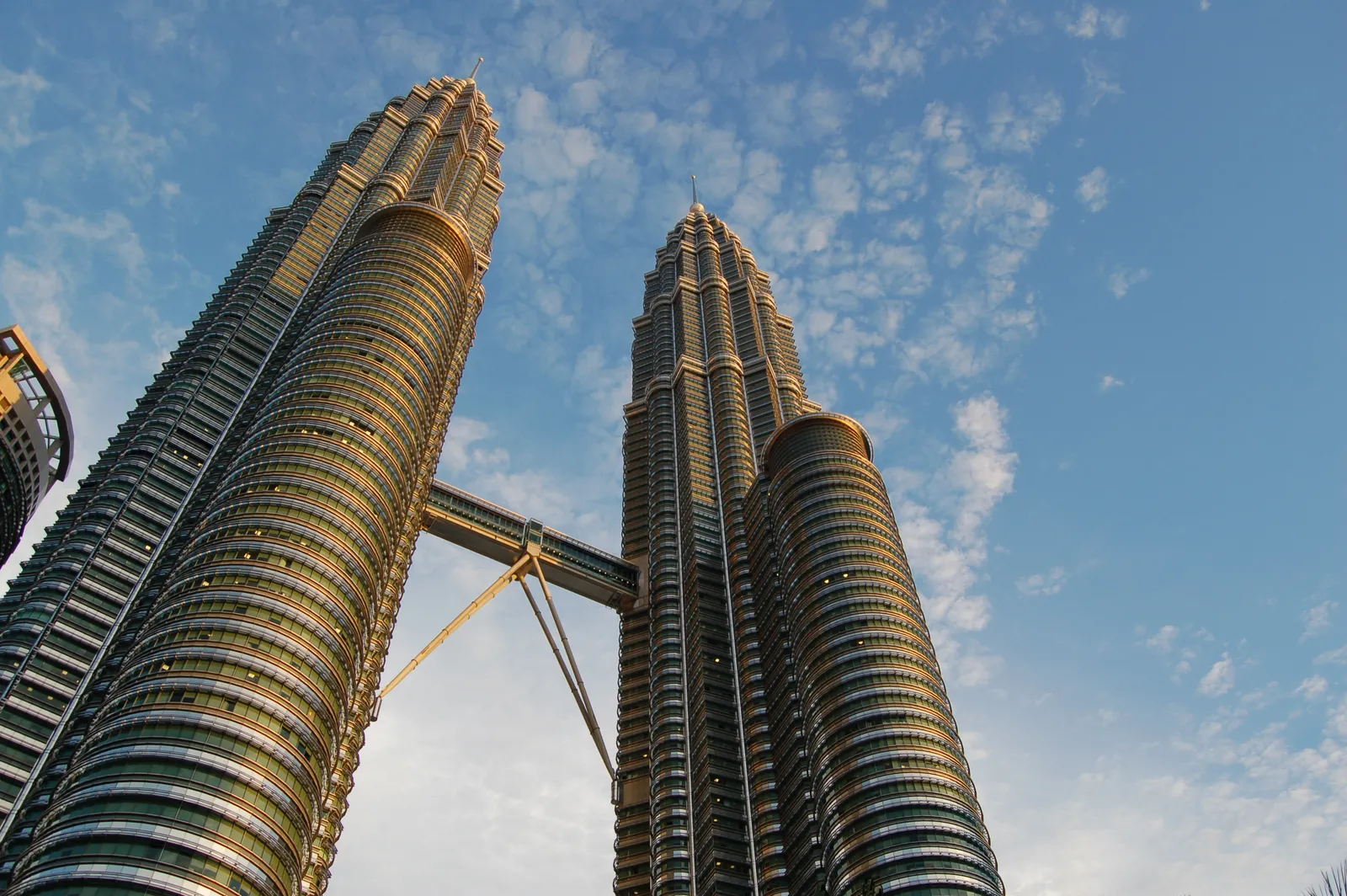 The Petronas Twin Towers seen from ground level against a blue sky, their stainless steel ribs tapering to twin spires with the two-storey skybridge braced between them partway up