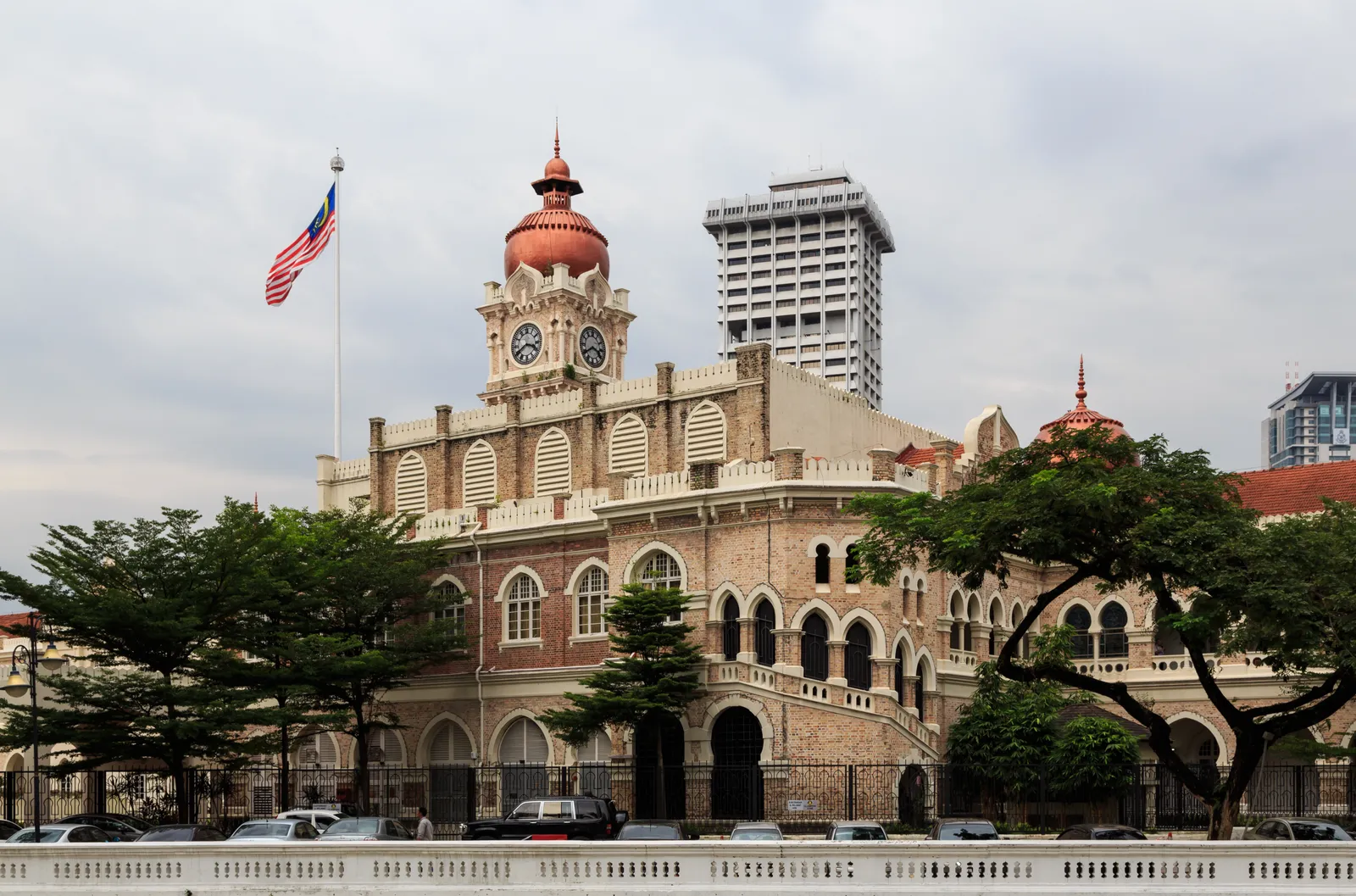 The Sultan Abdul Samad Building in Kuala Lumpur behind a row of trees, its copper-domed clock tower above a brick arcade, with the Malaysian flag flying from a pole in front