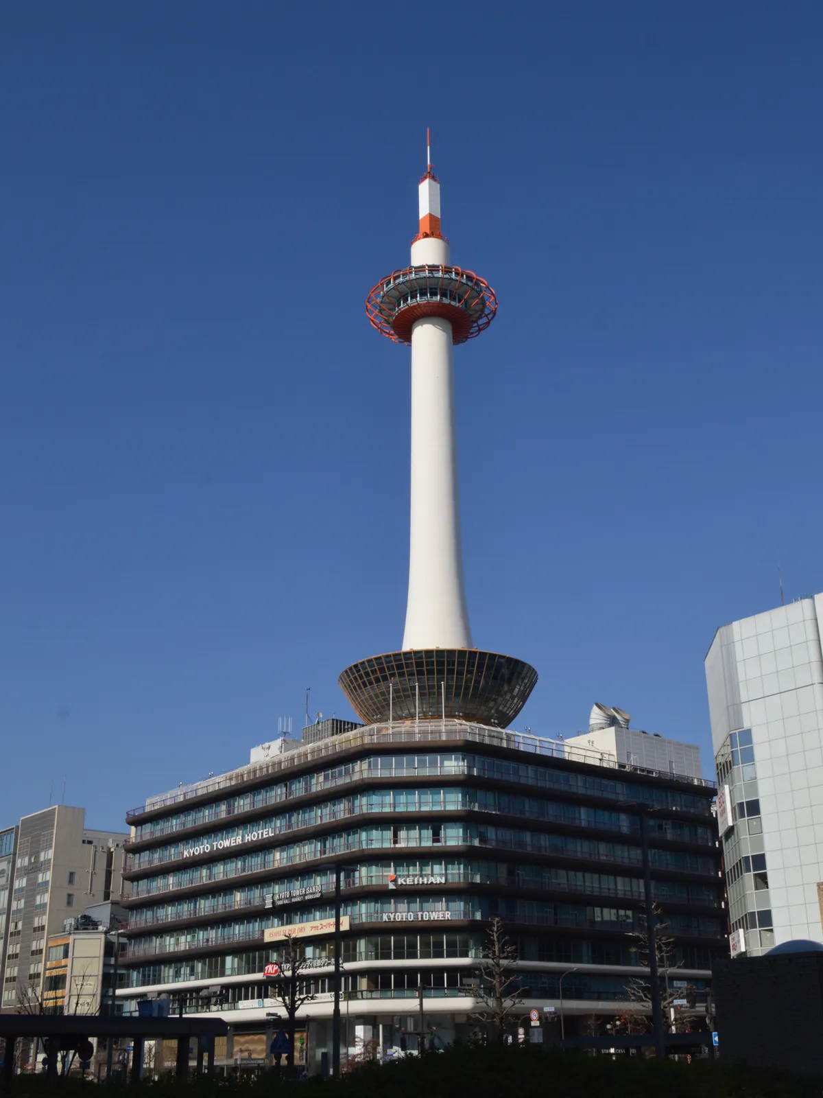 Kyoto Tower against a clear blue sky, a white tapering column with a red and white mast on top and a ringed observation deck partway up, rising from the flat-roofed building it stands on
