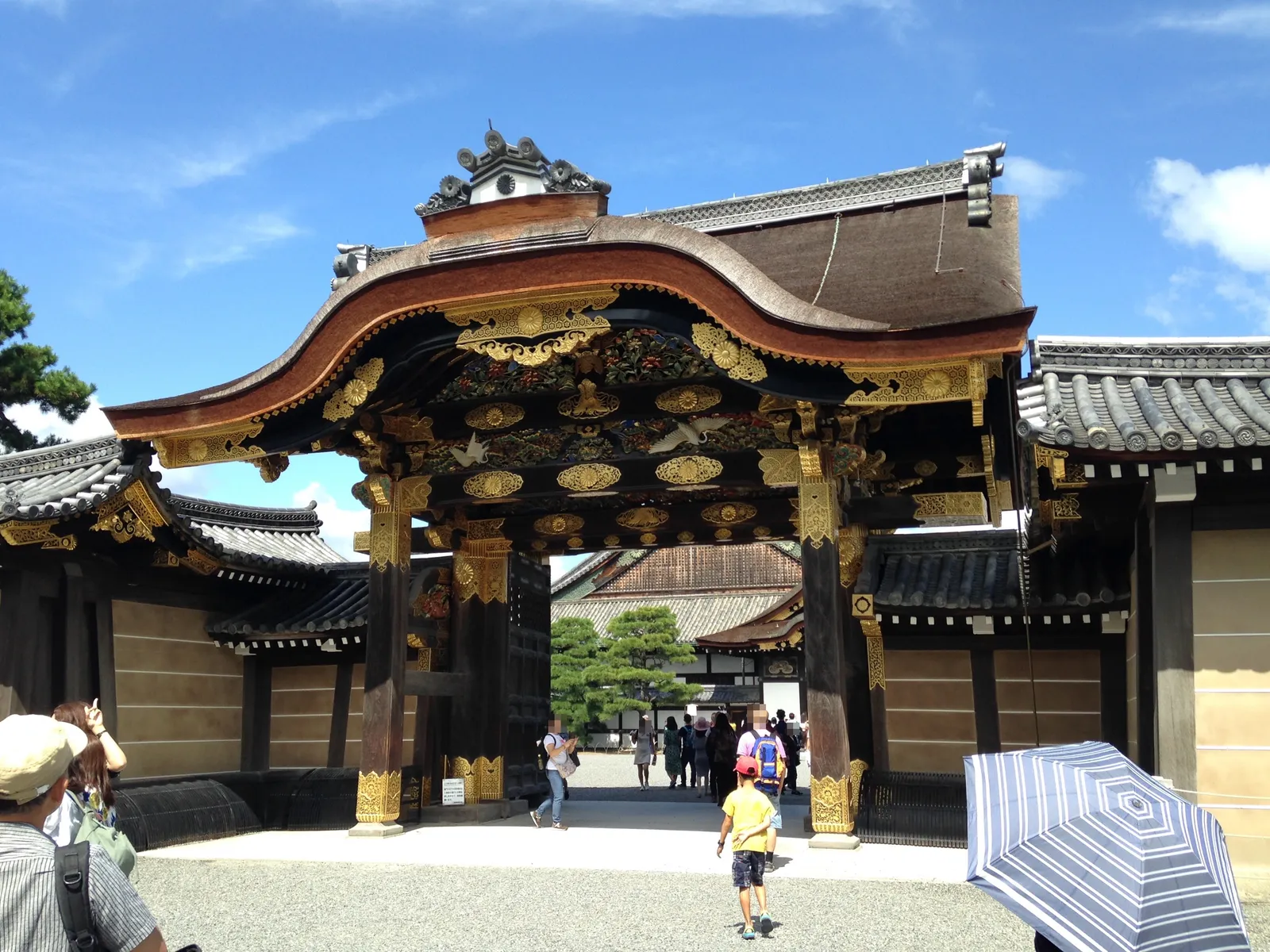 The Karamon gate at Nijo Castle in Kyoto, a curved cypress-bark roof over a gateway covered in gold leaf, carved birds and painted panels, with the palace roofs visible through the opening behind it