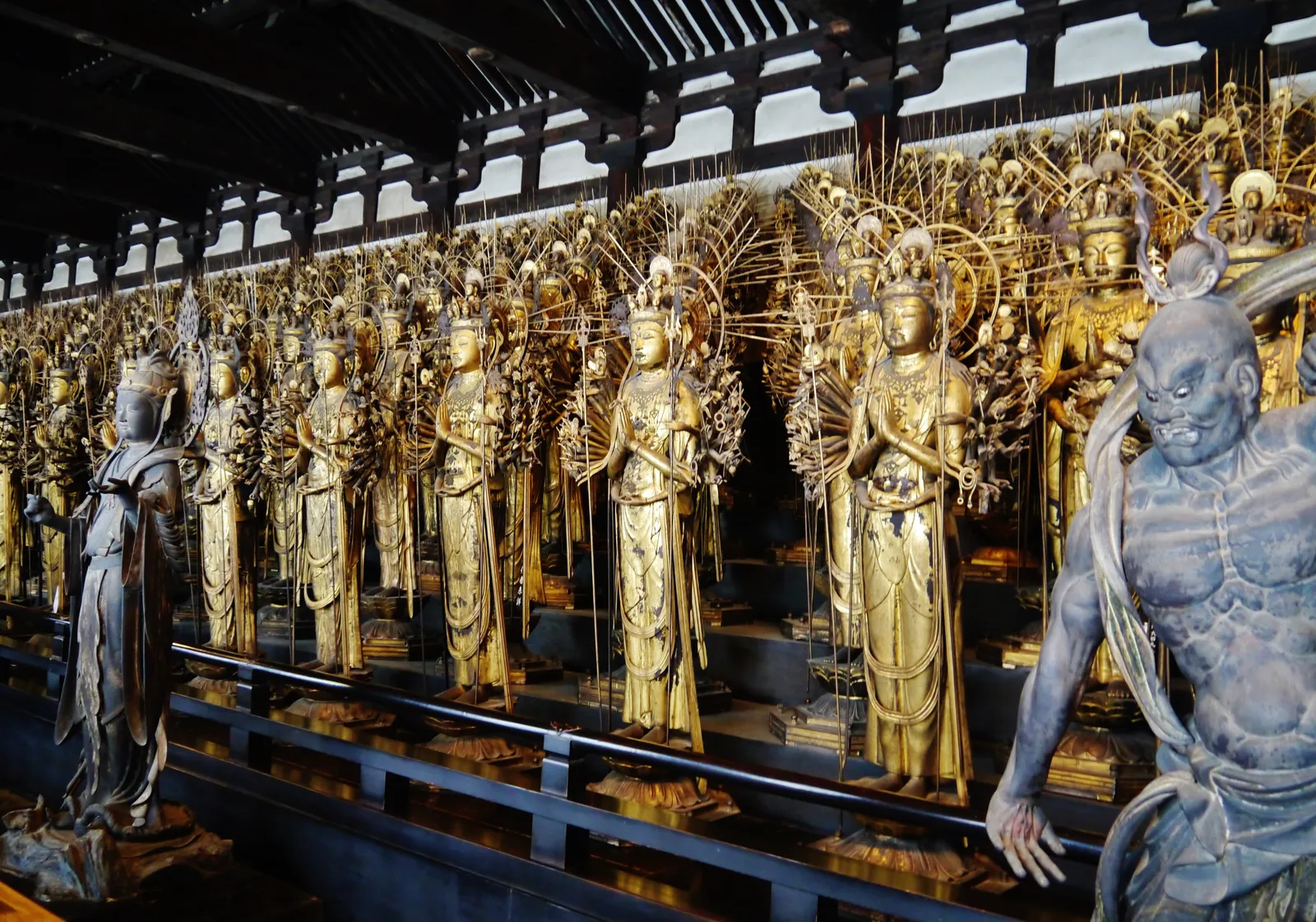 Rows of gilded, human-sized Kannon statues inside Sanjusangendo in Kyoto, each crowned with a fan of extra heads and arms, standing shoulder to shoulder in ranks that recede into the hall, with a dark wooden guardian figure in the foreground