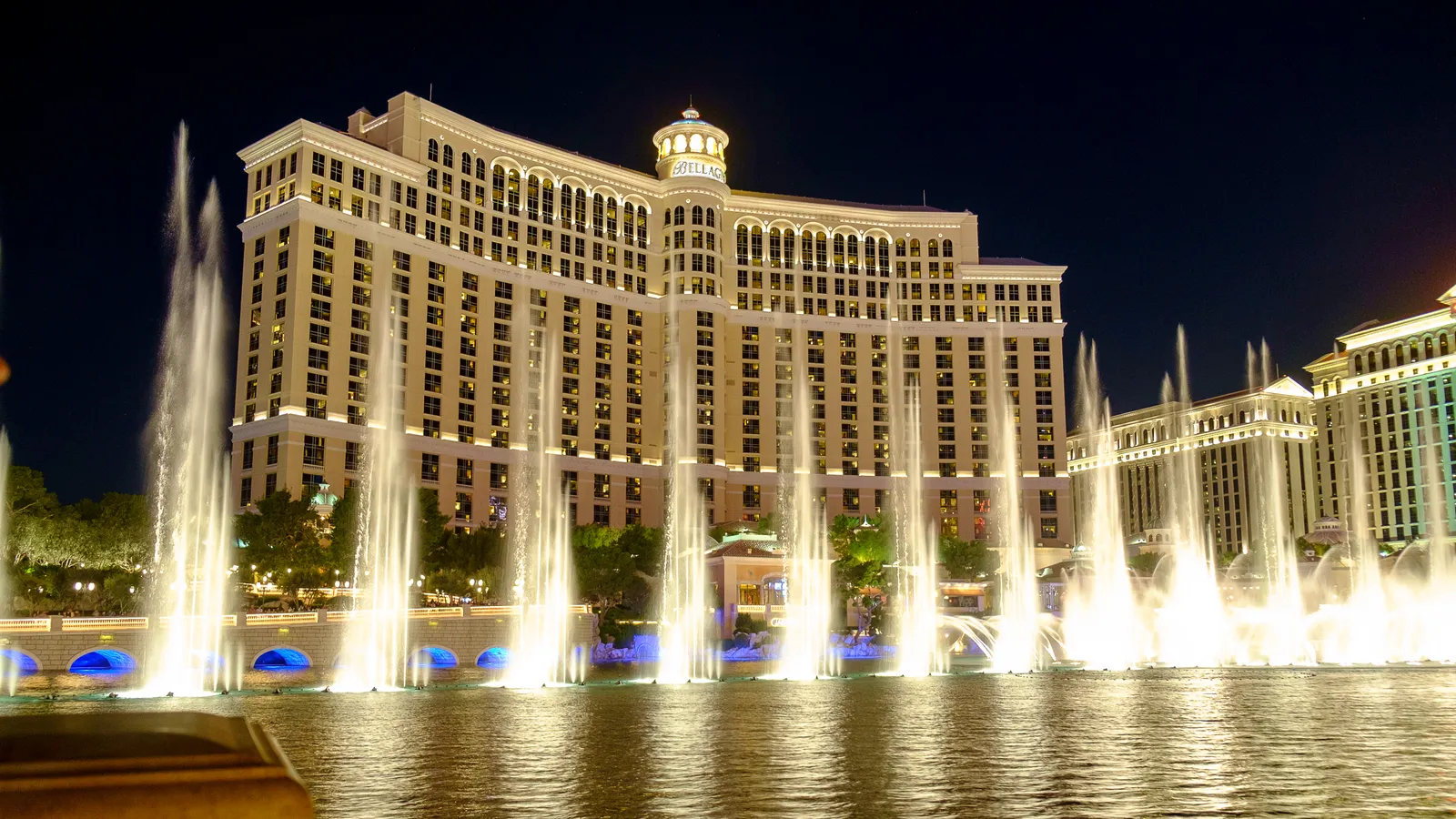 The Fountains of Bellagio at night, rows of lit water jets rising off the lake in front of the Bellagio's curved facade on the Las Vegas Strip