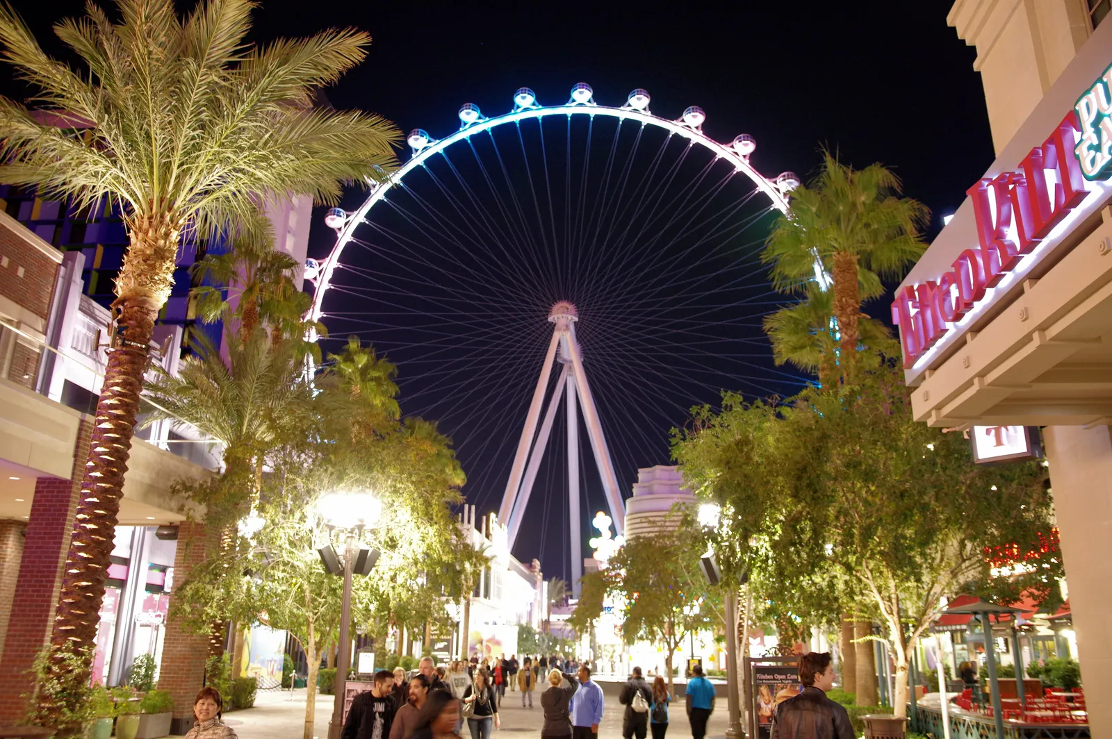The High Roller observation wheel lit at night at the end of the LINQ Promenade in Las Vegas, with palm trees and people walking the lane below it