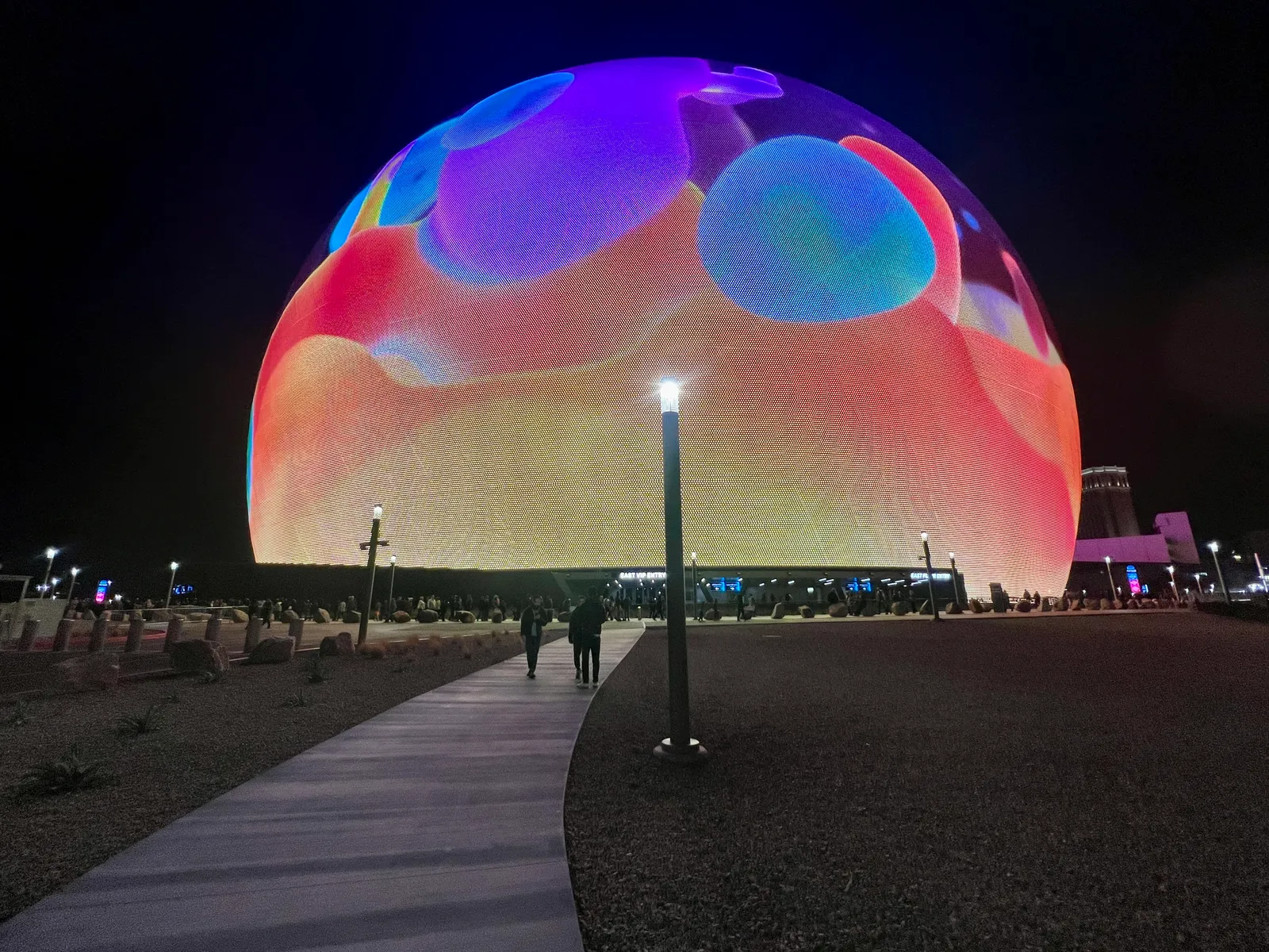 Sphere in Las Vegas at night, its whole curved exterior lit in pink, orange and blue, with a lamp-lit footpath and people walking toward the entrance