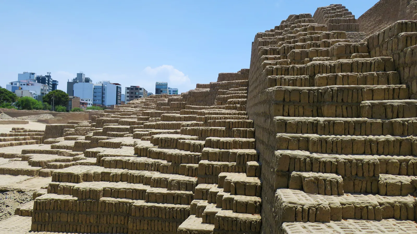 The stepped adobe walls of Huaca Pucllana in Miraflores, with thousands of small clay bricks stood upright side by side and Lima apartment towers rising behind the site