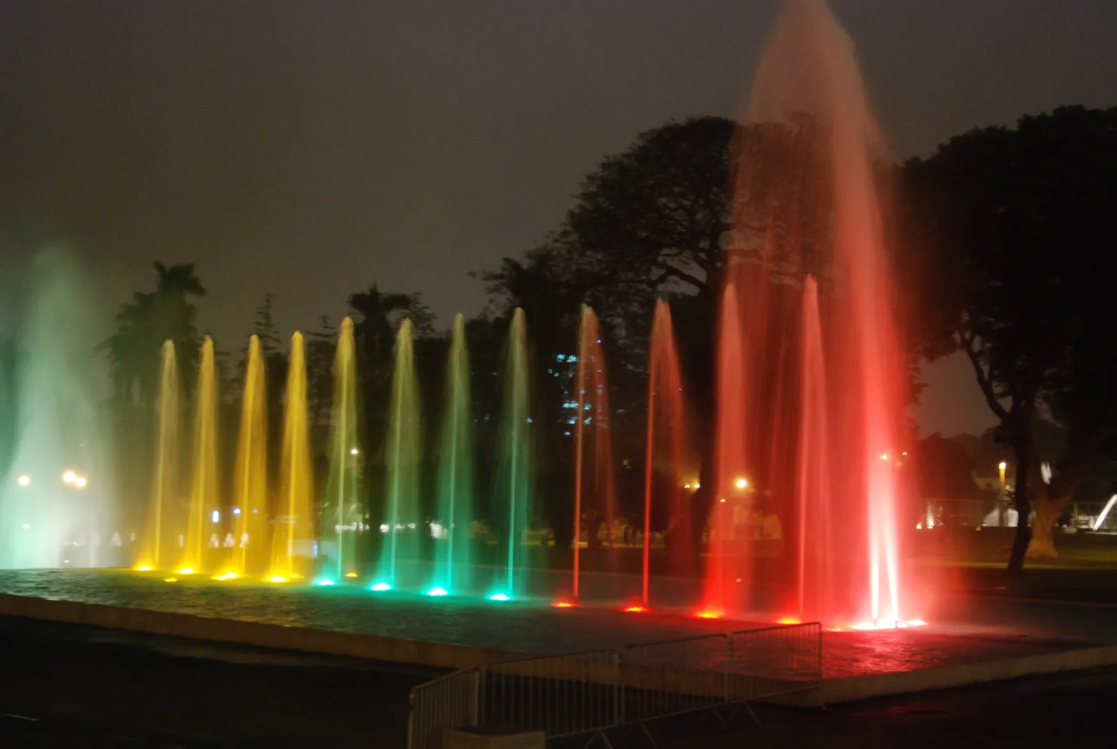 A row of fountain jets in Lima's Parque de la Reserva lit green, yellow and red against a dark evening sky