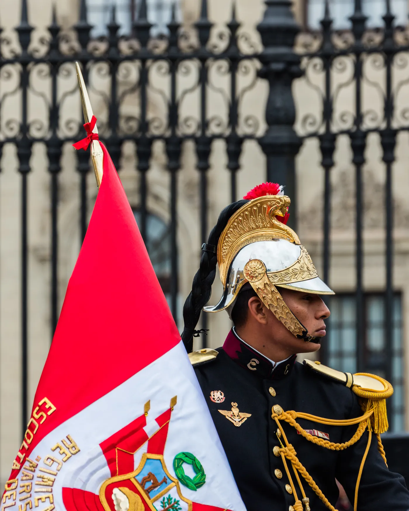 A soldier of Peru's presidential escort in a plumed gold helmet and dark dress uniform carrying the national flag outside the iron railings of the Government Palace in Lima