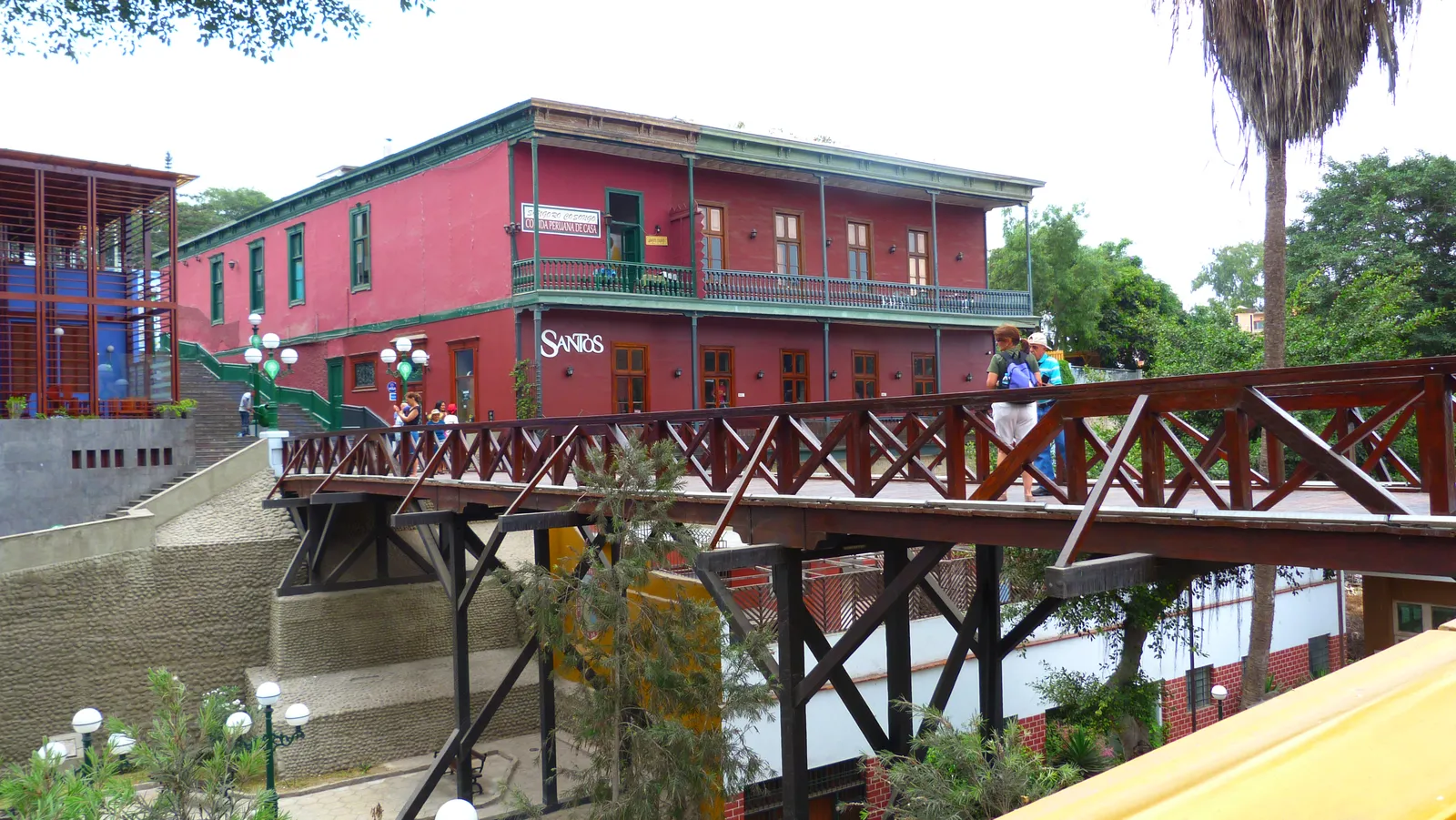 The wooden Puente de los Suspiros in Barranco, Lima, with people crossing it and a red two-storey house with a balcony behind