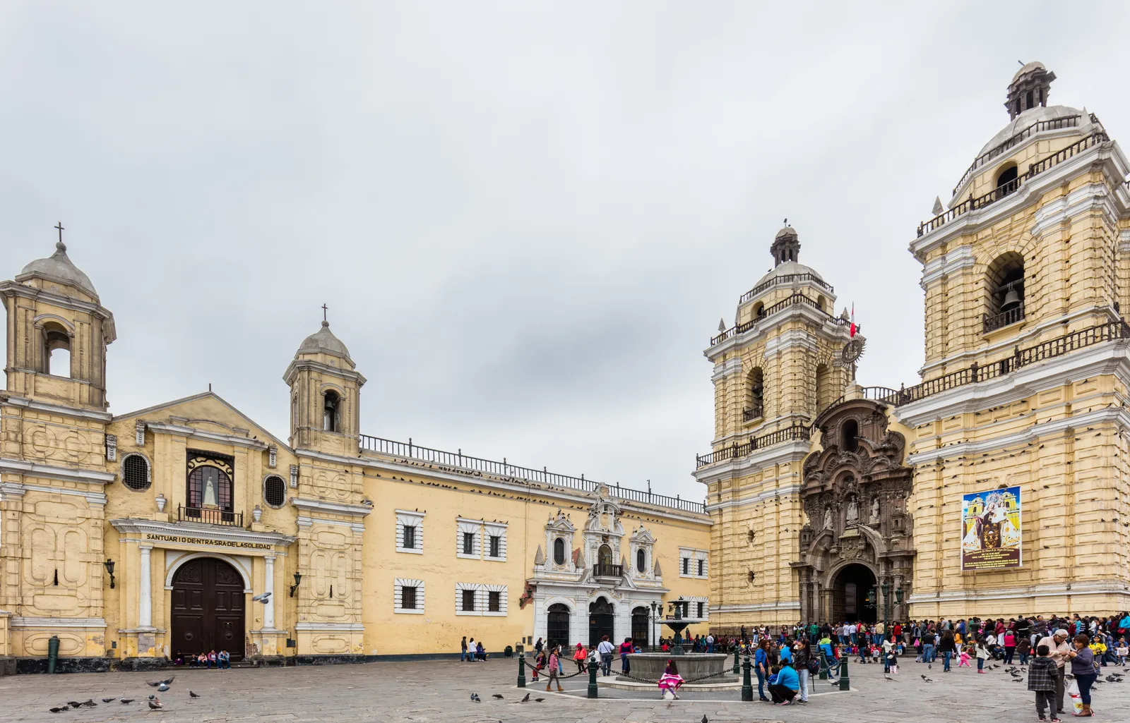 The yellow baroque facade of the Basilica and Convent of San Francisco in Lima, with its two bell towers and the walled forecourt full of visitors and pigeons