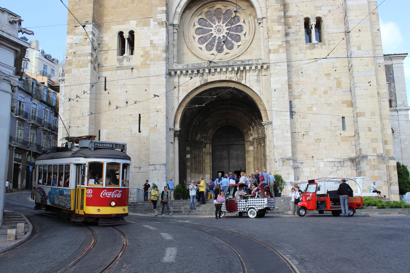The twin Romanesque towers and rose window of Lisbon Cathedral, with a yellow number 28 tram passing in front of the entrance