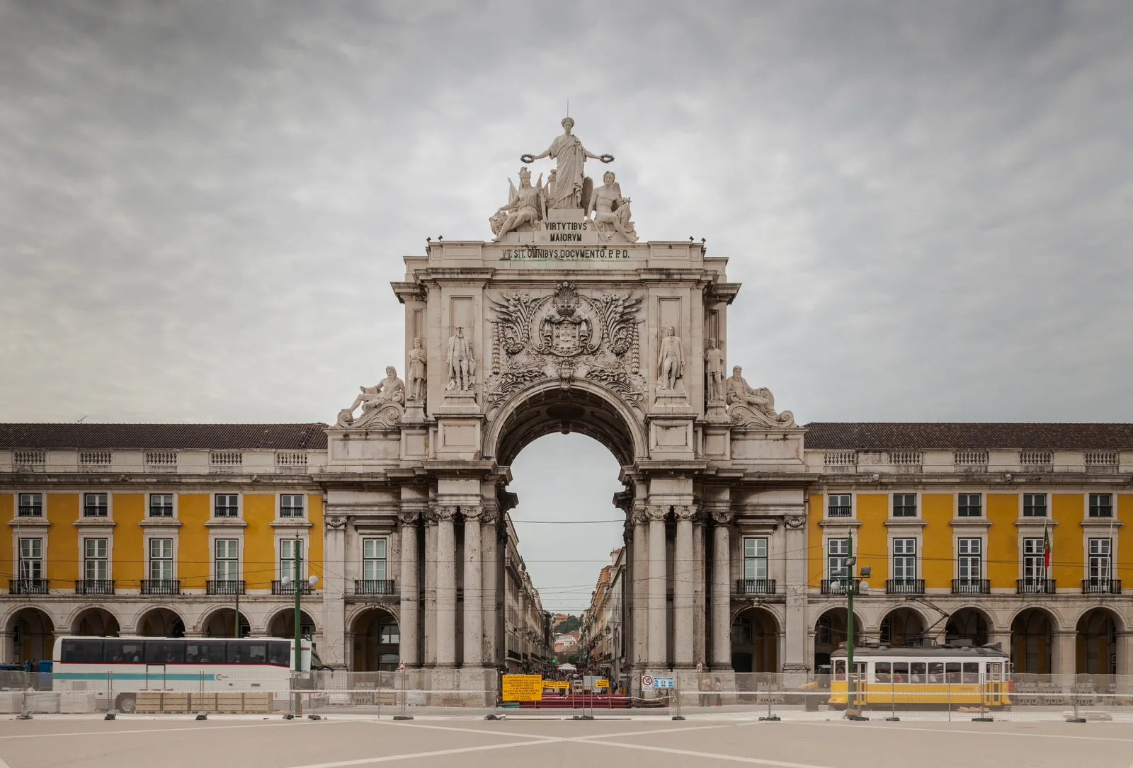 The triumphal arch at the top of Rua Augusta in Lisbon, framed by the yellow arcaded buildings of Praça do Comércio, with a yellow tram passing beneath it