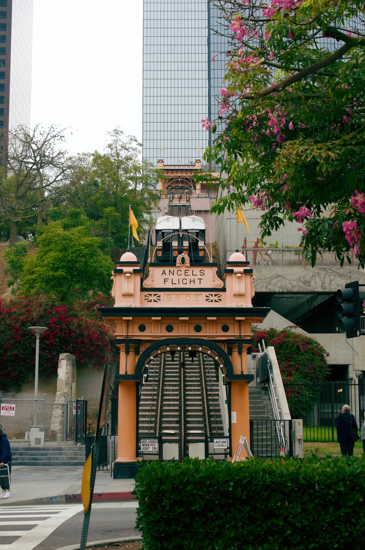 The lower station of Angels Flight in Los Angeles, an orange and black archway lettered with the railway's name, with the funicular track and its wooden car climbing the slope behind it towards a glass tower