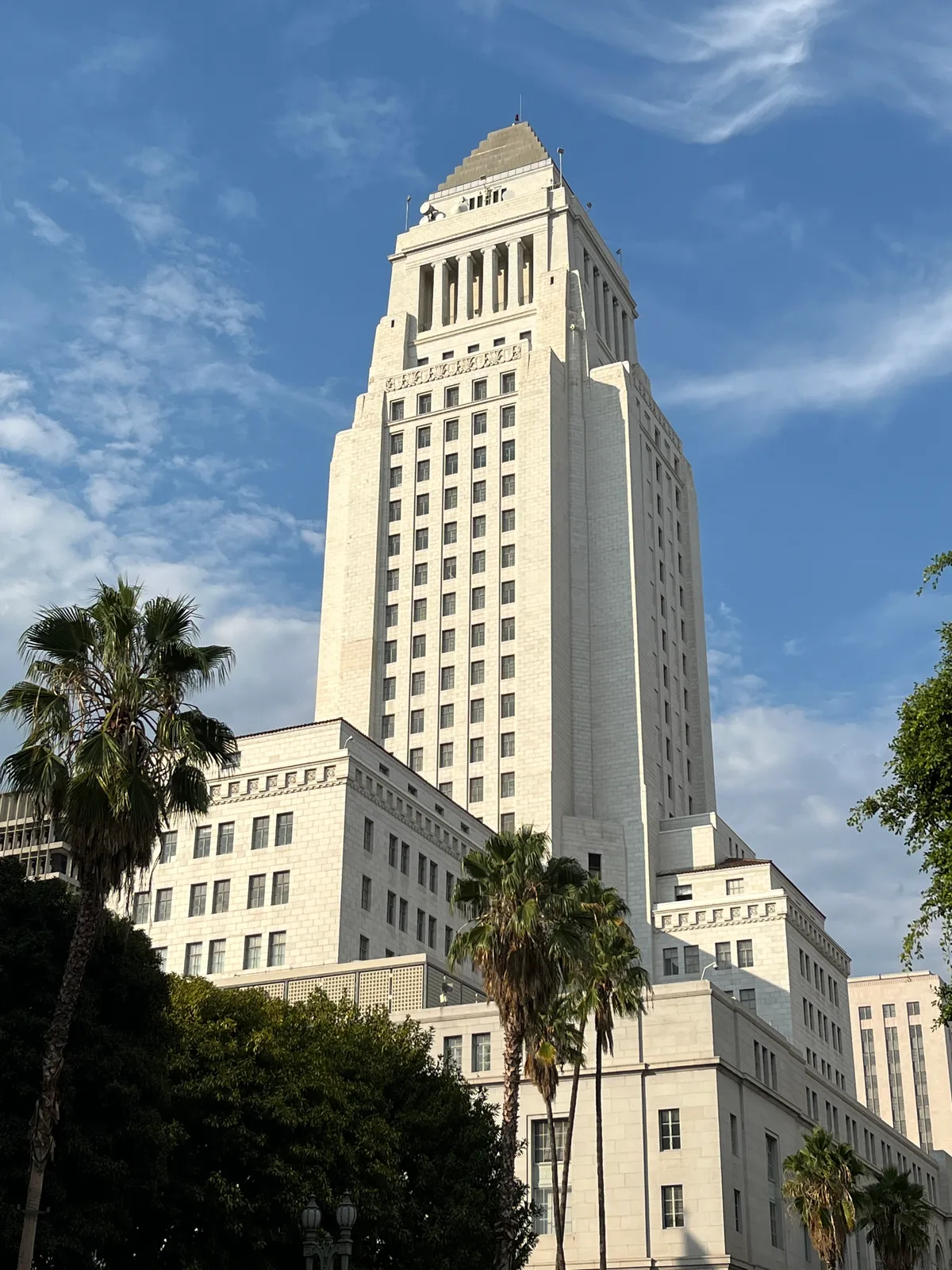Los Angeles City Hall from below, a white stepped tower rising above a broad stone base, with palm trees along the street in front of it and a clear blue sky behind