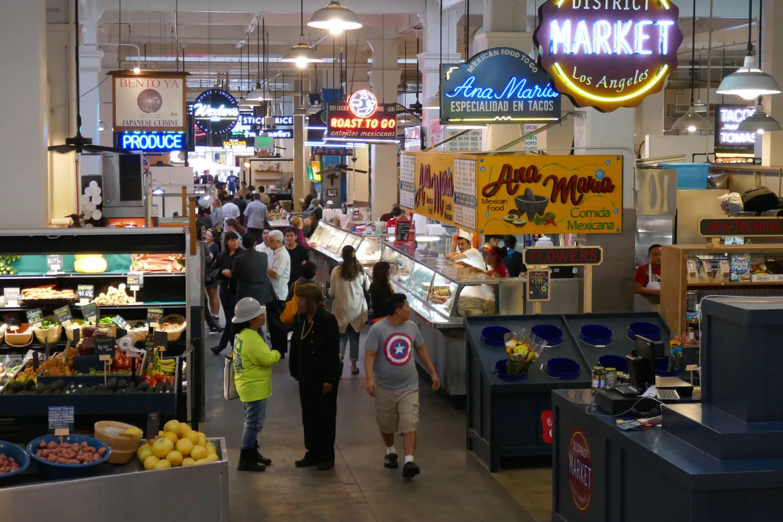 The interior of Grand Central Market in Los Angeles, a wide aisle between food counters under hanging lamps, with neon vendor signs above the stalls and crates of fruit and vegetables in the foreground