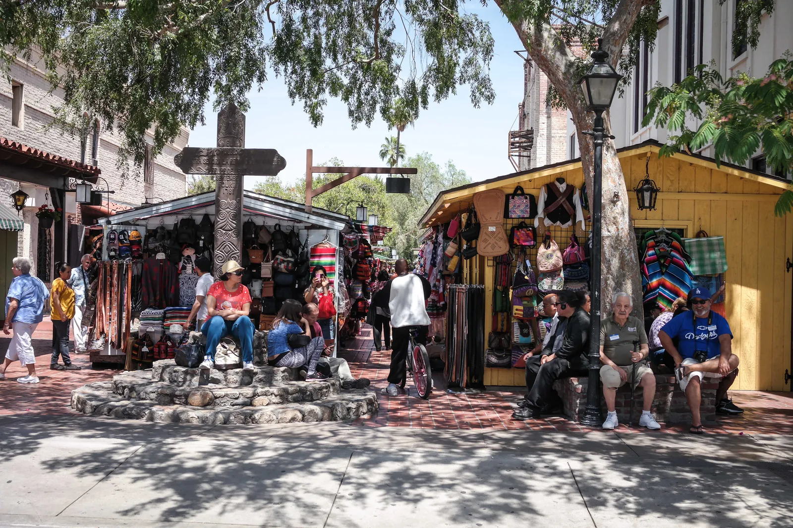 Olvera Street in Los Angeles, a brick-paved pedestrian lane lined with covered market stalls hung with blankets, bags and guitars, shaded by trees, with people sitting on a low stone wall