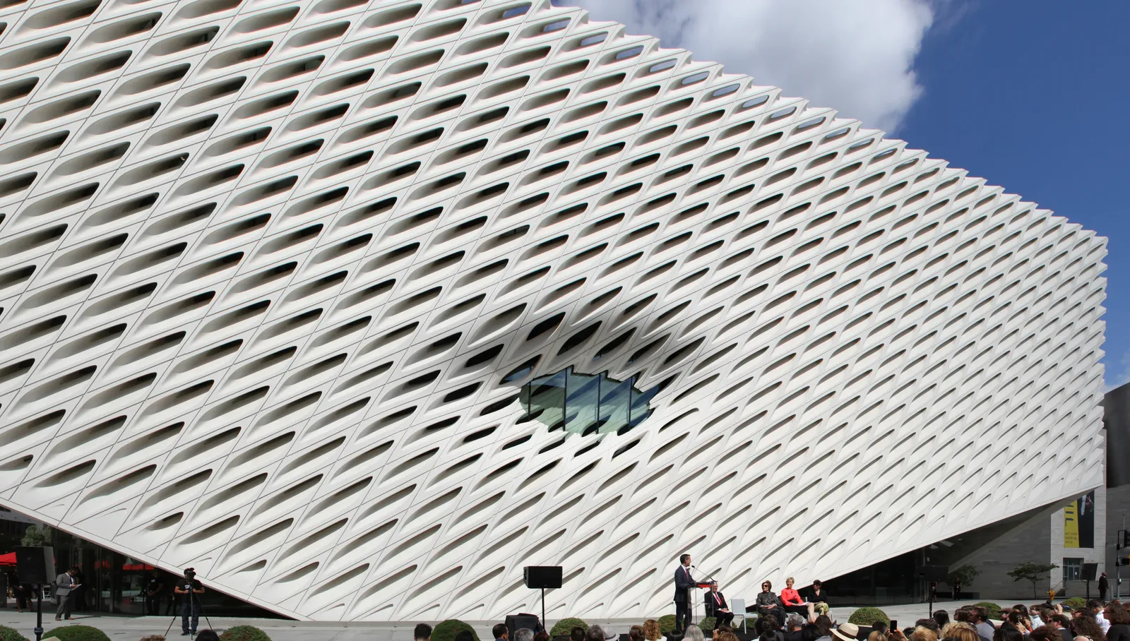 The Broad museum in Los Angeles, a long white facade of angled honeycomb cells wrapping the building, with a single large window scooped out of the middle of it and people standing at street level below