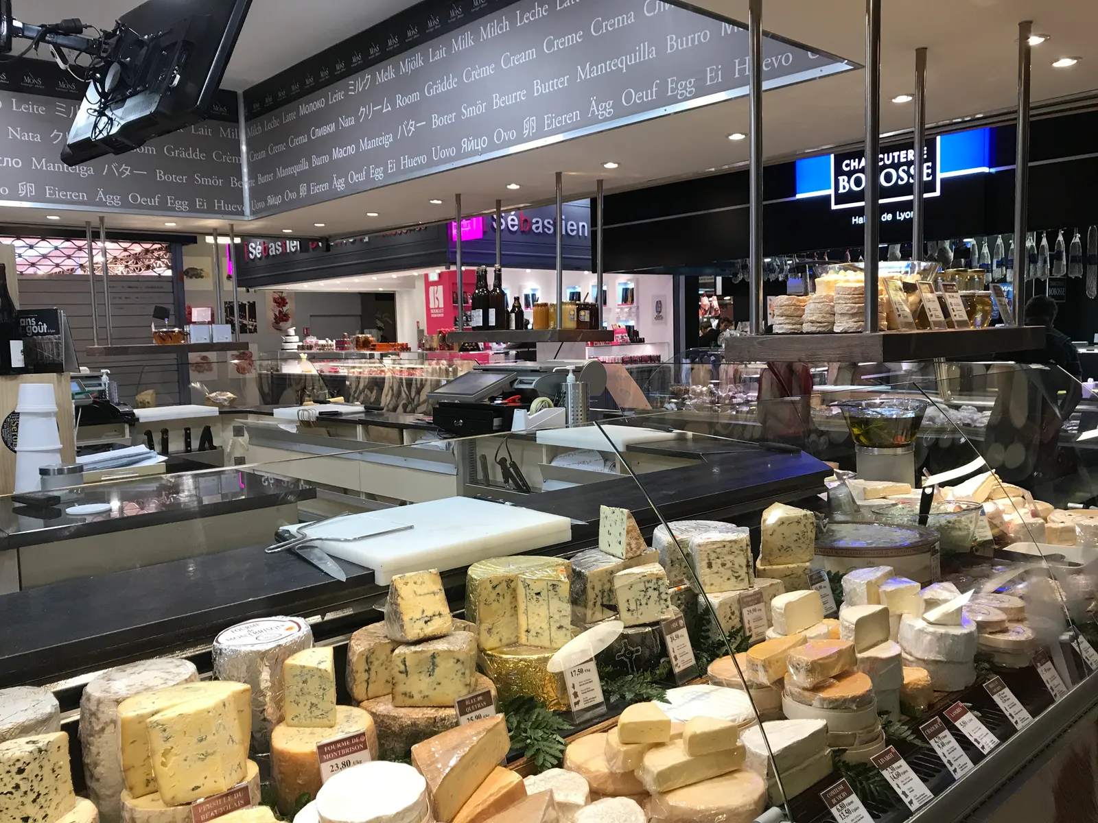 A cheese counter inside Les Halles de Lyon Paul Bocuse, dozens of whole cheeses stacked along a refrigerated display with price cards, and stall signs above