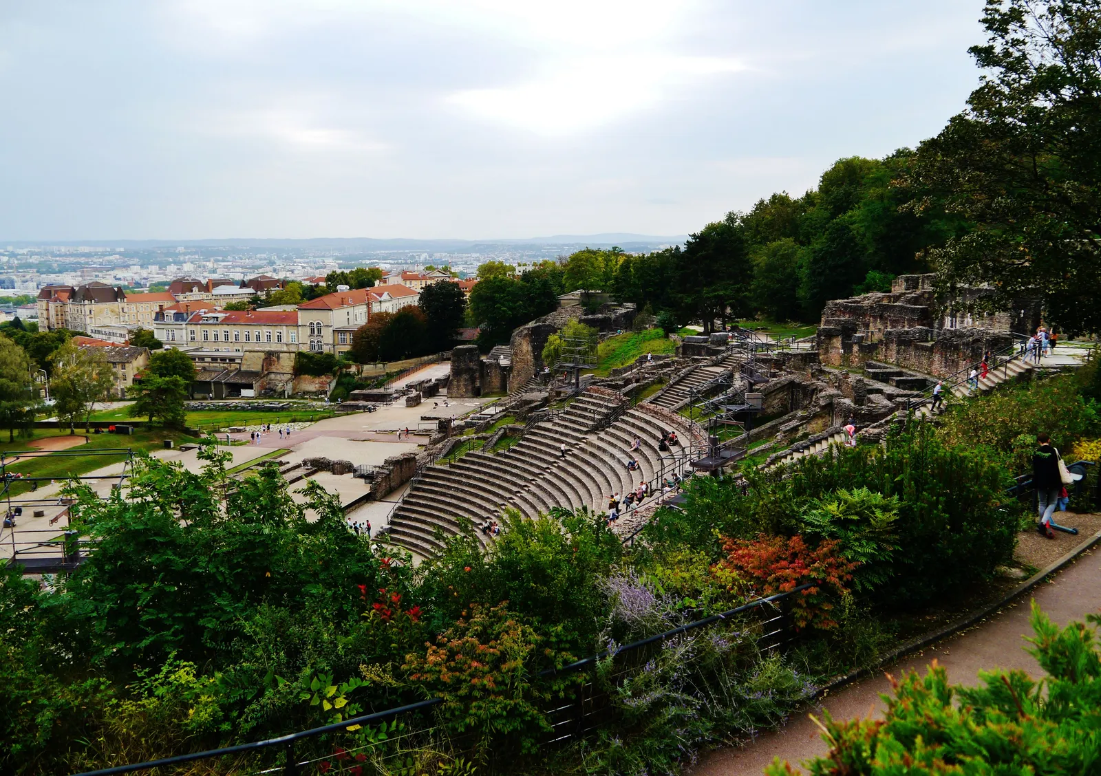 The Roman theatre on the Fourvière hillside in Lyon seen from above, its semicircle of stone seating tiers cut into the slope with the city rooftops behind