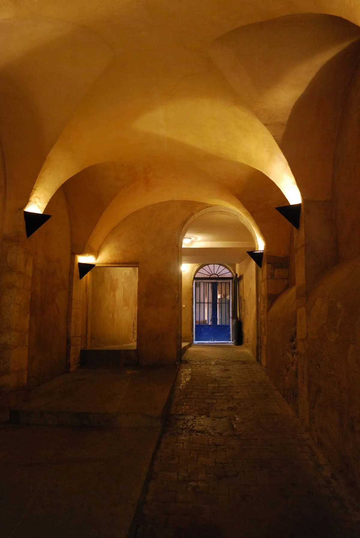The inside of a covered passage in Vieux Lyon, a vaulted stone ceiling lit by wall lamps above a worn flagstone floor, with an archway and a blue door at the far end