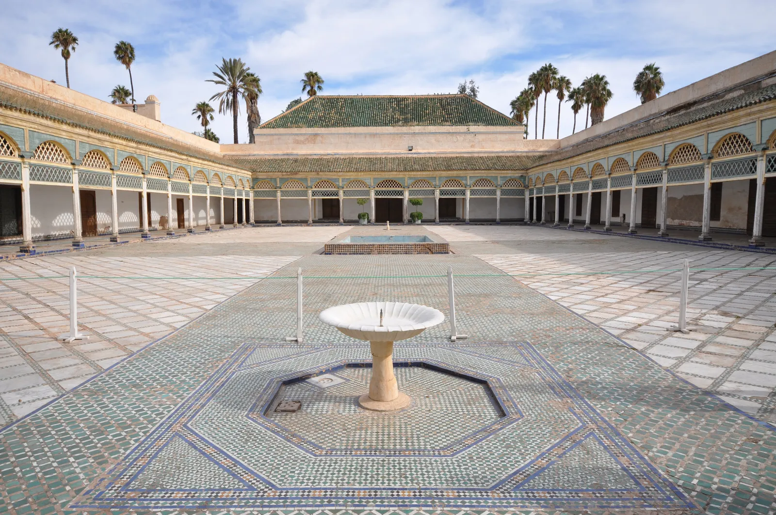 The Grand Courtyard of the Bahia Palace in Marrakech, a wide marble floor with a white fountain on a tiled star in the centre, surrounded by a painted wooden gallery under green tiled roofs and palm trees
