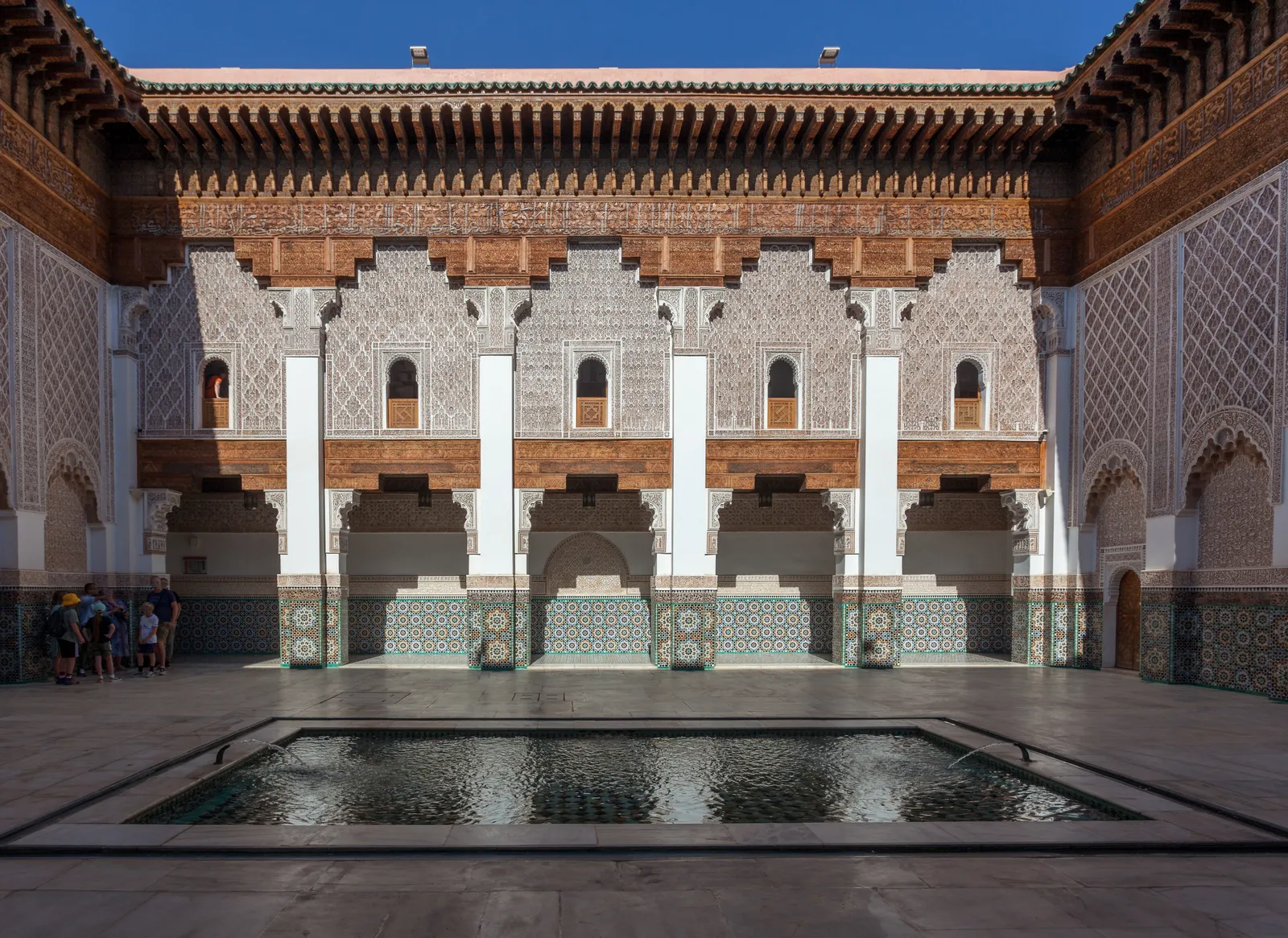 The courtyard of the Ben Youssef Madrasa in Marrakech, a long rectangular pool in front of an arcade of zellige-tiled pillars, carved stucco walls and a row of small windows from the student rooms under a carved cedar cornice