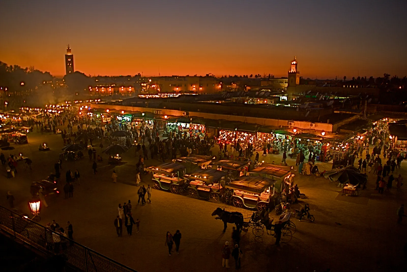 Jemaa el-Fnaa in Marrakech just after sunset, the food stalls lit up and smoking in the middle of the square, crowds and horse-drawn carriages in the foreground and the Koutoubia minaret silhouetted against an orange sky