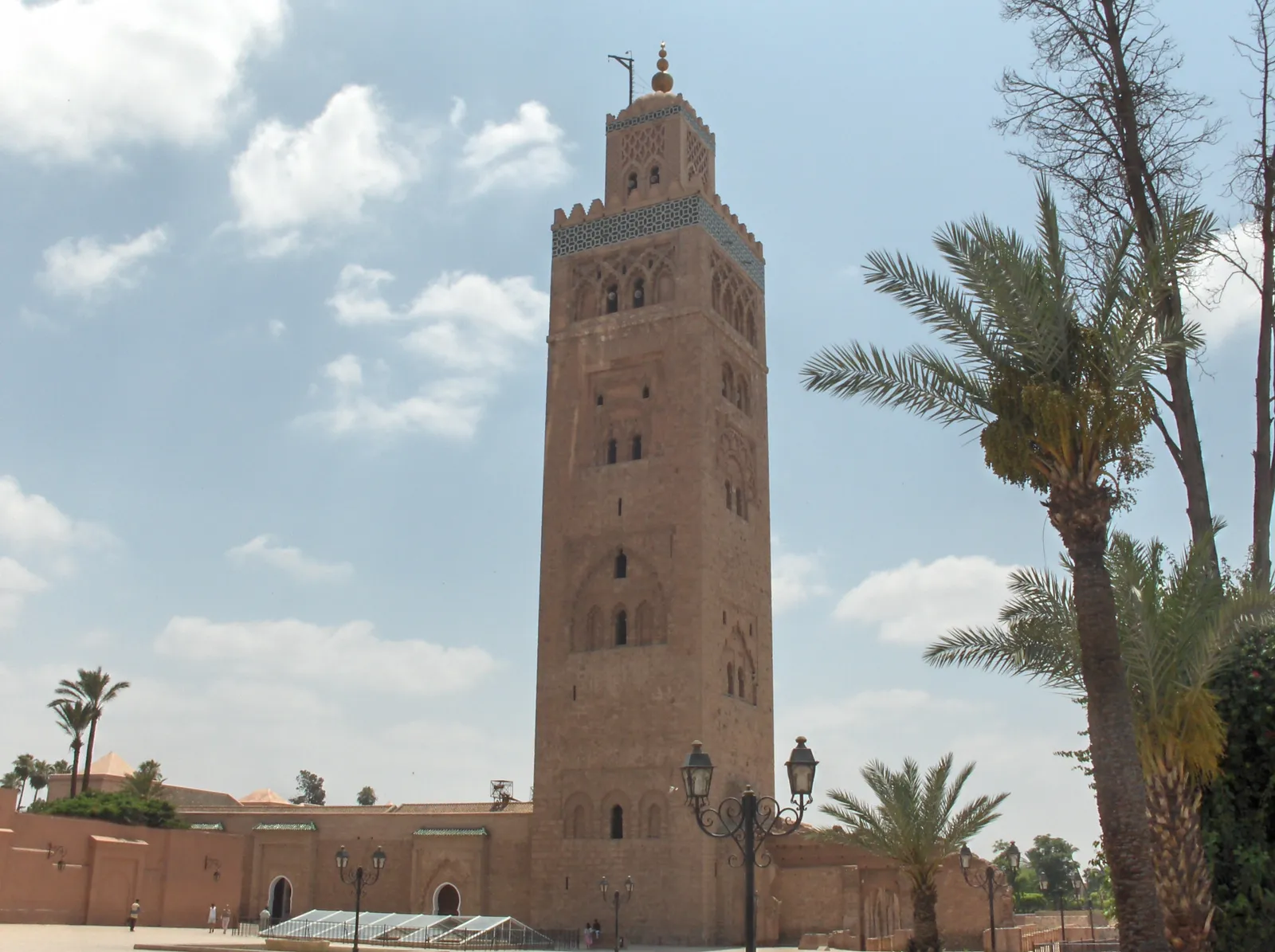 The Koutoubia minaret in Marrakech, a tall square tower of pinkish stone with carved window arches and a band of turquoise tile near the top, standing above the mosque's low walls with palm trees and a street lamp in front