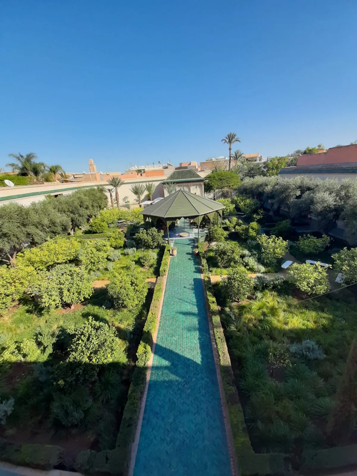 Le Jardin Secret in Marrakech seen from its tower, a green-tiled water channel running straight through dense planted beds to a pavilion with a green pyramid roof, medina rooftops and palms beyond the garden wall