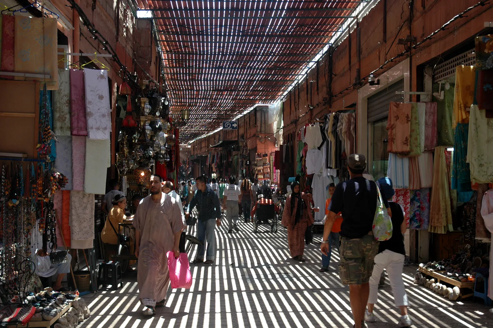 Souk Semmarine in Marrakech under its slatted reed canopy, stripes of sunlight falling across the lane, with scarves, lamps and slippers hanging from the shopfronts on both sides and shoppers walking through
