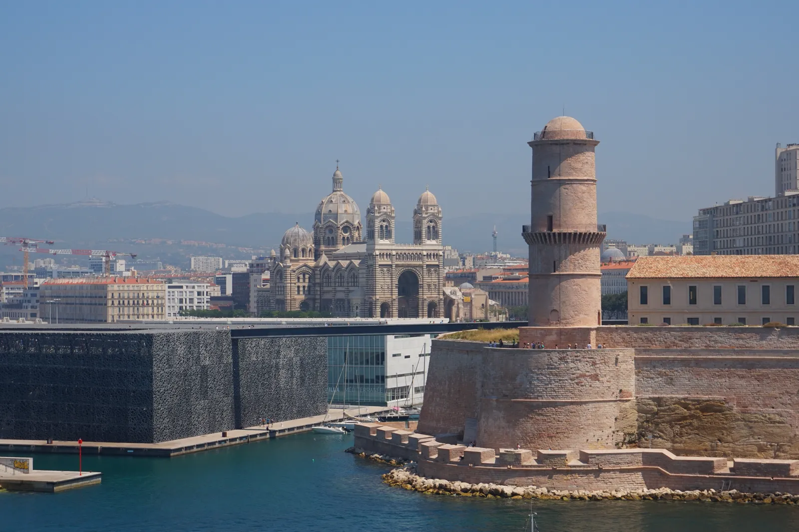 Fort Saint-Jean's stone tower and ramparts at the mouth of Marseille's Old Port, with the MuCEM's black lattice-clad building beside it and Marseille Cathedral's striped domes visible across the water