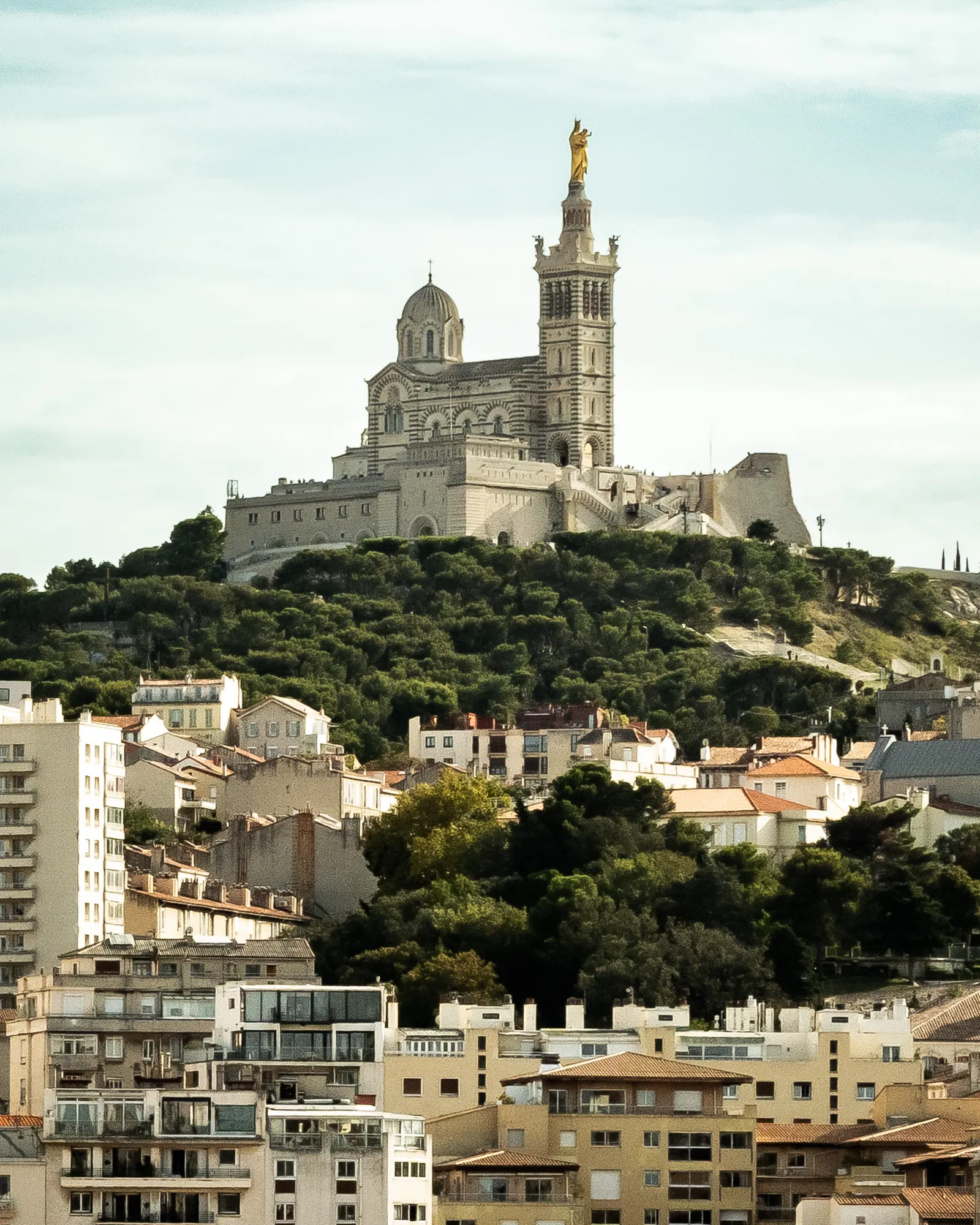 Notre-Dame de la Garde basilica topped with its gilded statue of the Virgin Mary, standing on a wooded hilltop above Marseille's rooftops