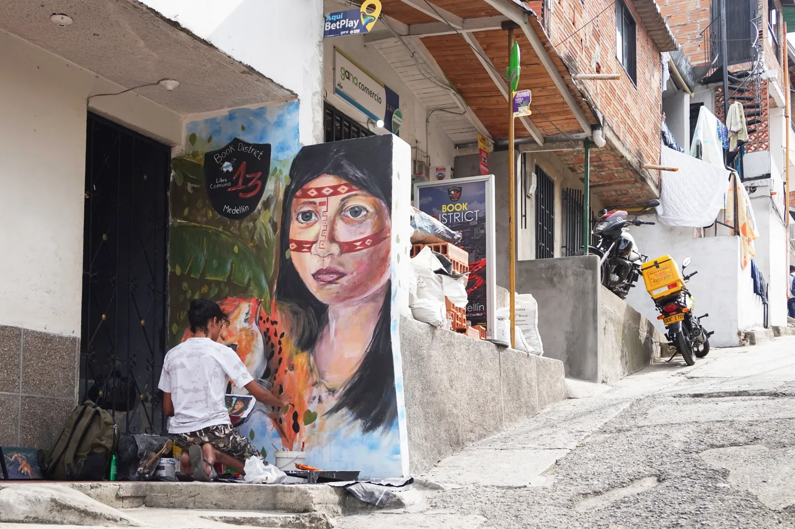 A street artist painting a portrait mural on a wall in Comuna 13, Medellín, with motorbikes parked along the narrow street