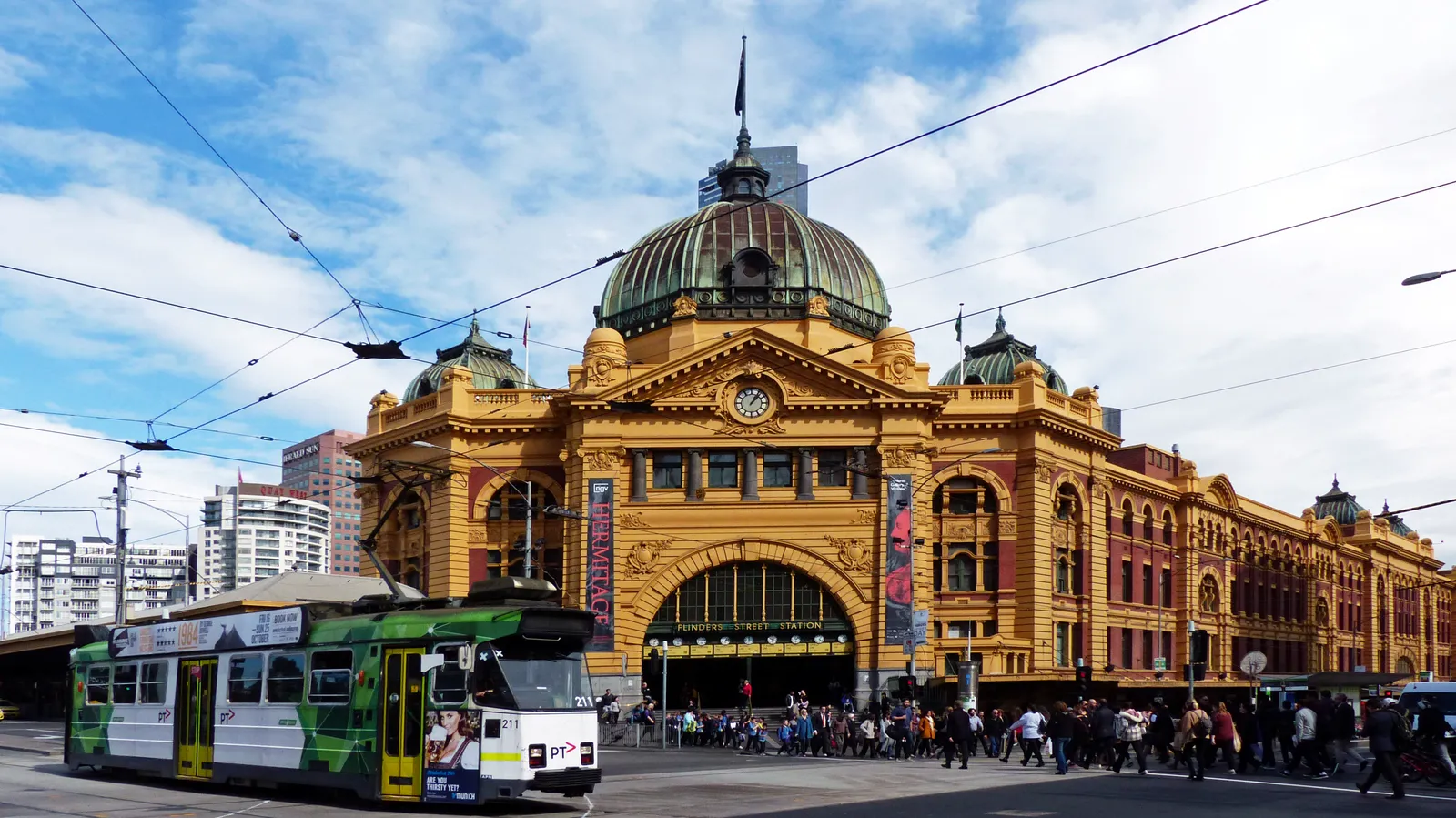 The yellow stucco frontage and green copper dome of Flinders Street Station in Melbourne, with a tram passing in front of the main entrance