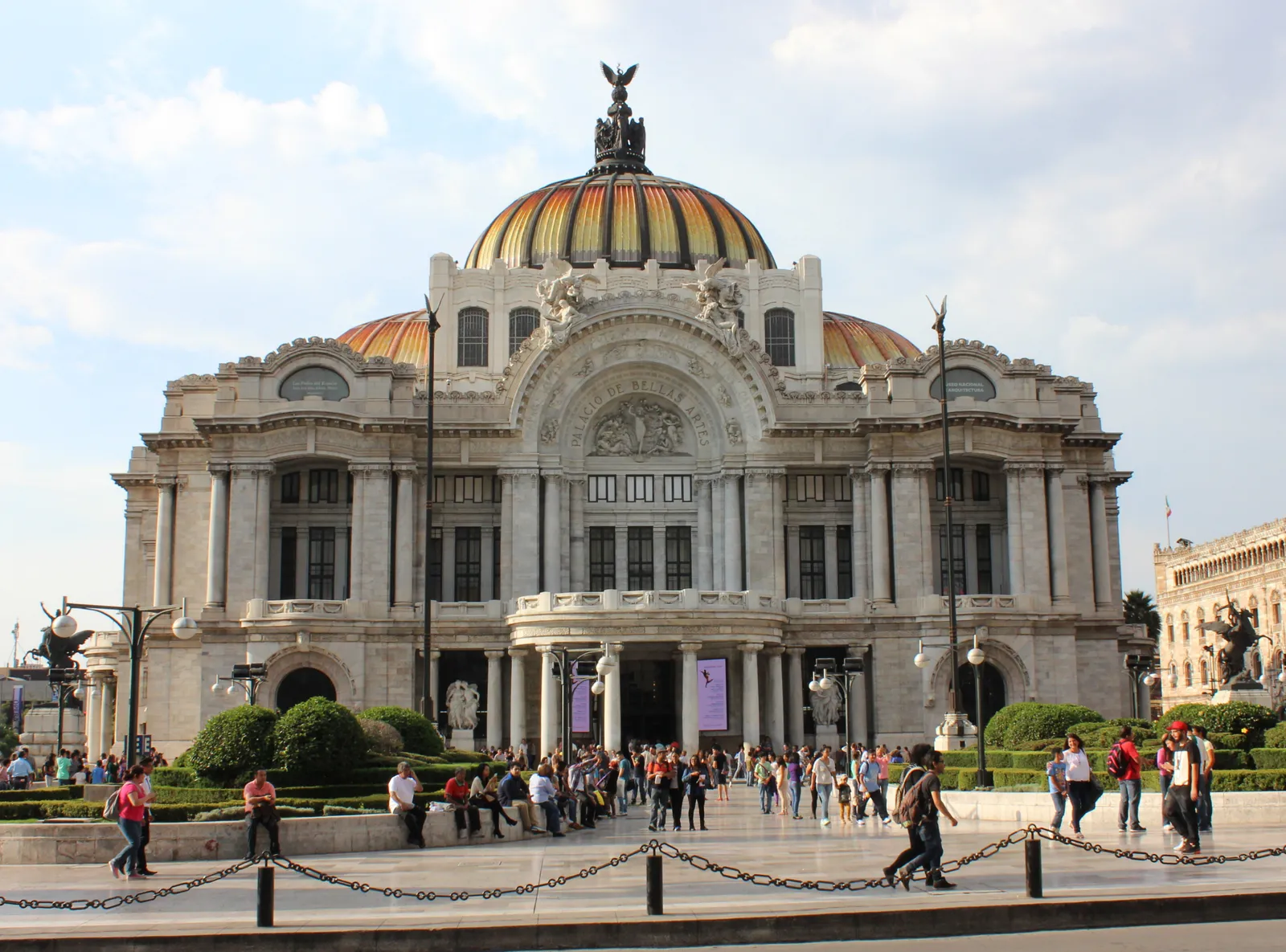 The white marble front of the Palacio de Bellas Artes in Mexico City, with its orange and yellow tiled dome and a crowd of people sitting on the steps below the entrance