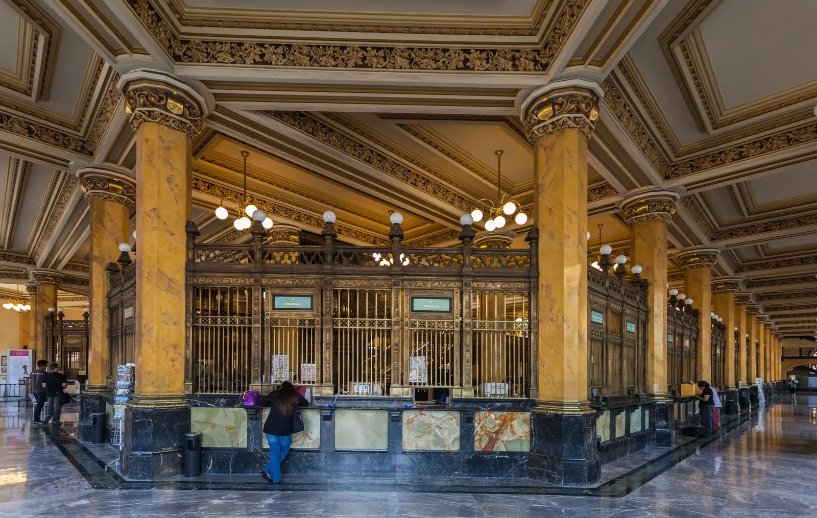 The main hall of Mexico City's Palacio Postal, lined with marble columns and gilded ironwork counters under a coffered ceiling, with a customer being served at one of the windows
