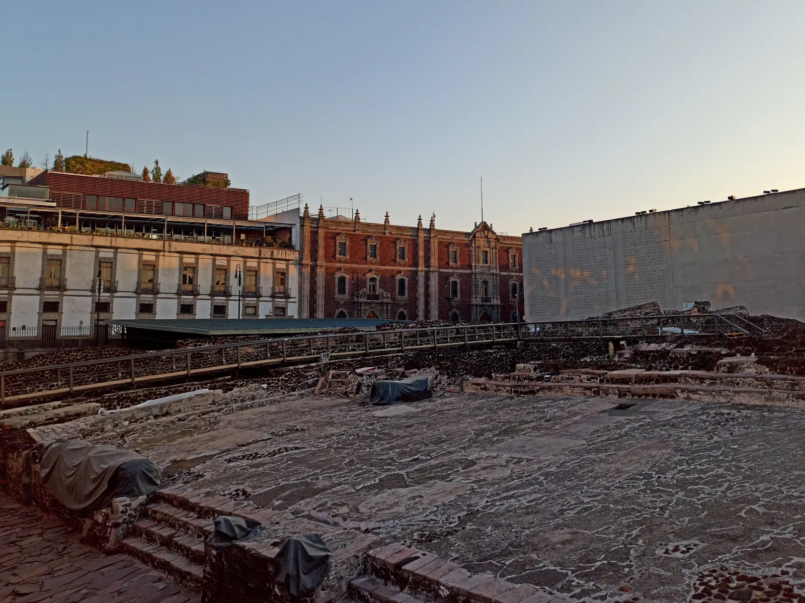 The excavated stone platforms and stairways of the Templo Mayor in Mexico City, sunk below street level with a colonial-era building and modern walls rising directly behind them