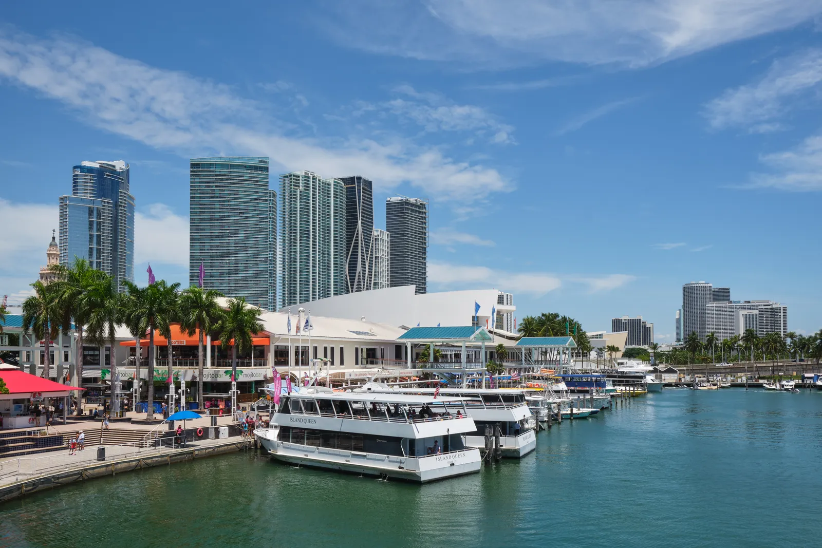 White sightseeing boats moored in the marina at Bayside Marketplace in Miami, with the marketplace's low buildings, palm trees and downtown towers behind them